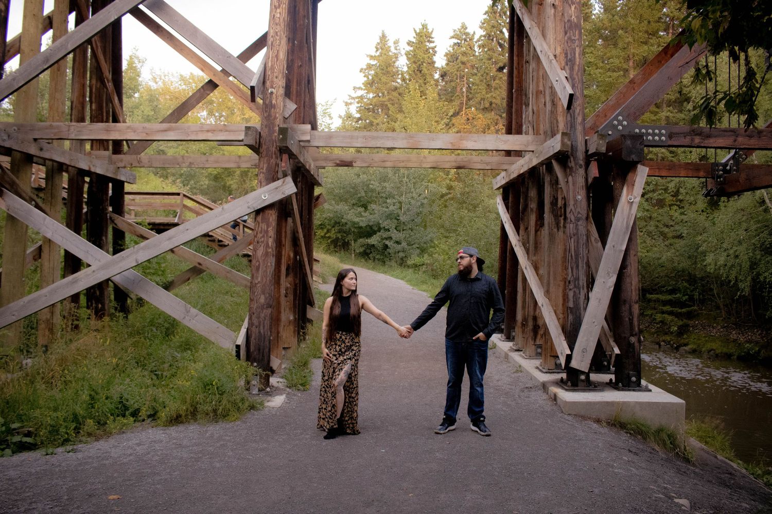 A couple holding hands while walking under a rustic wooden bridge structure.