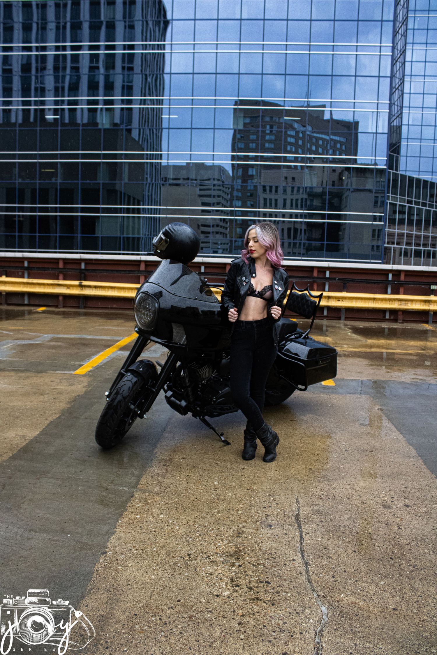 Modern black motorcycle parked on wet concrete in front of glass office buildings with a person posing beside it.