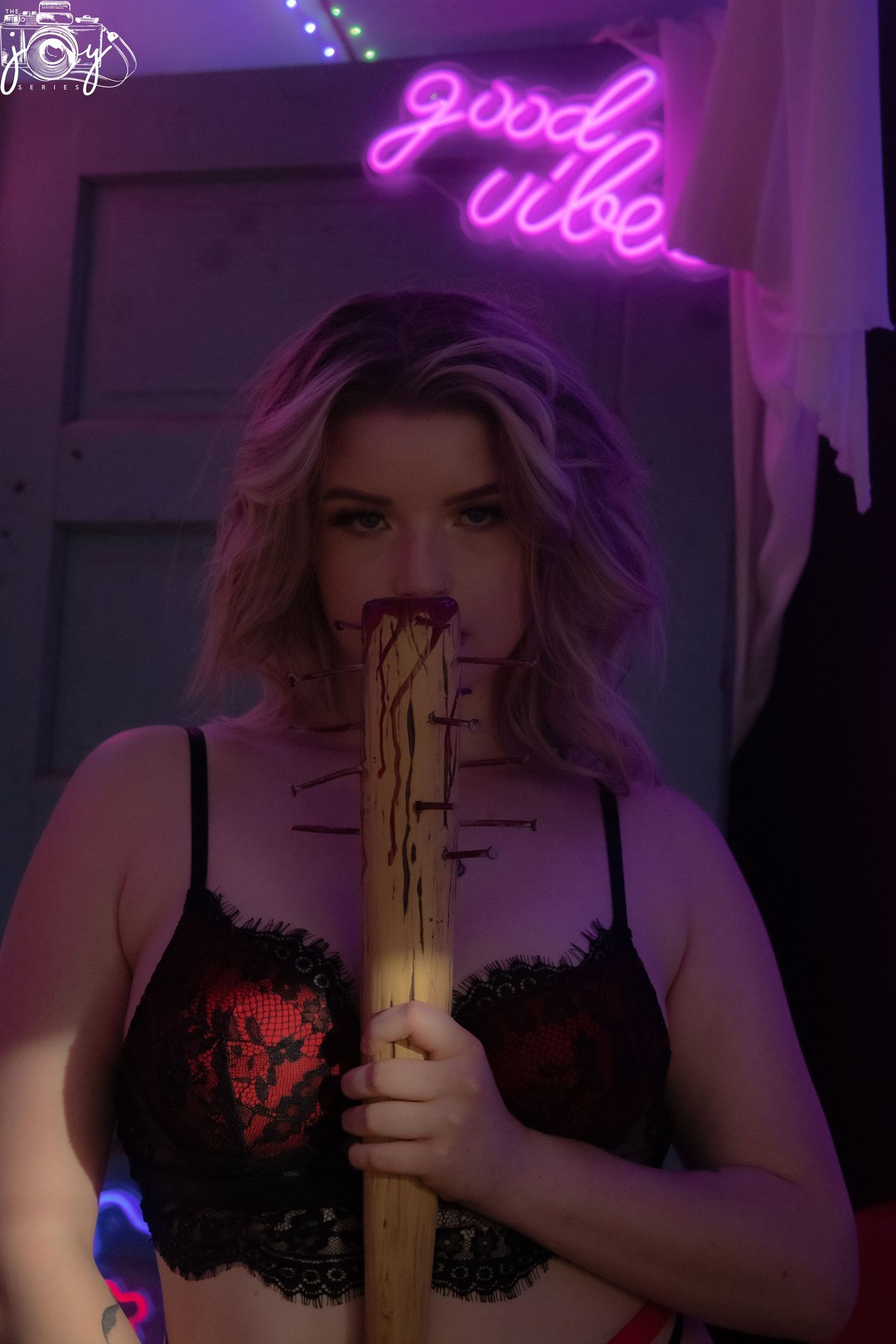 A moody boudoir photo with purple neon lighting showing 'good vibe' text and someone holding a baseball bat.