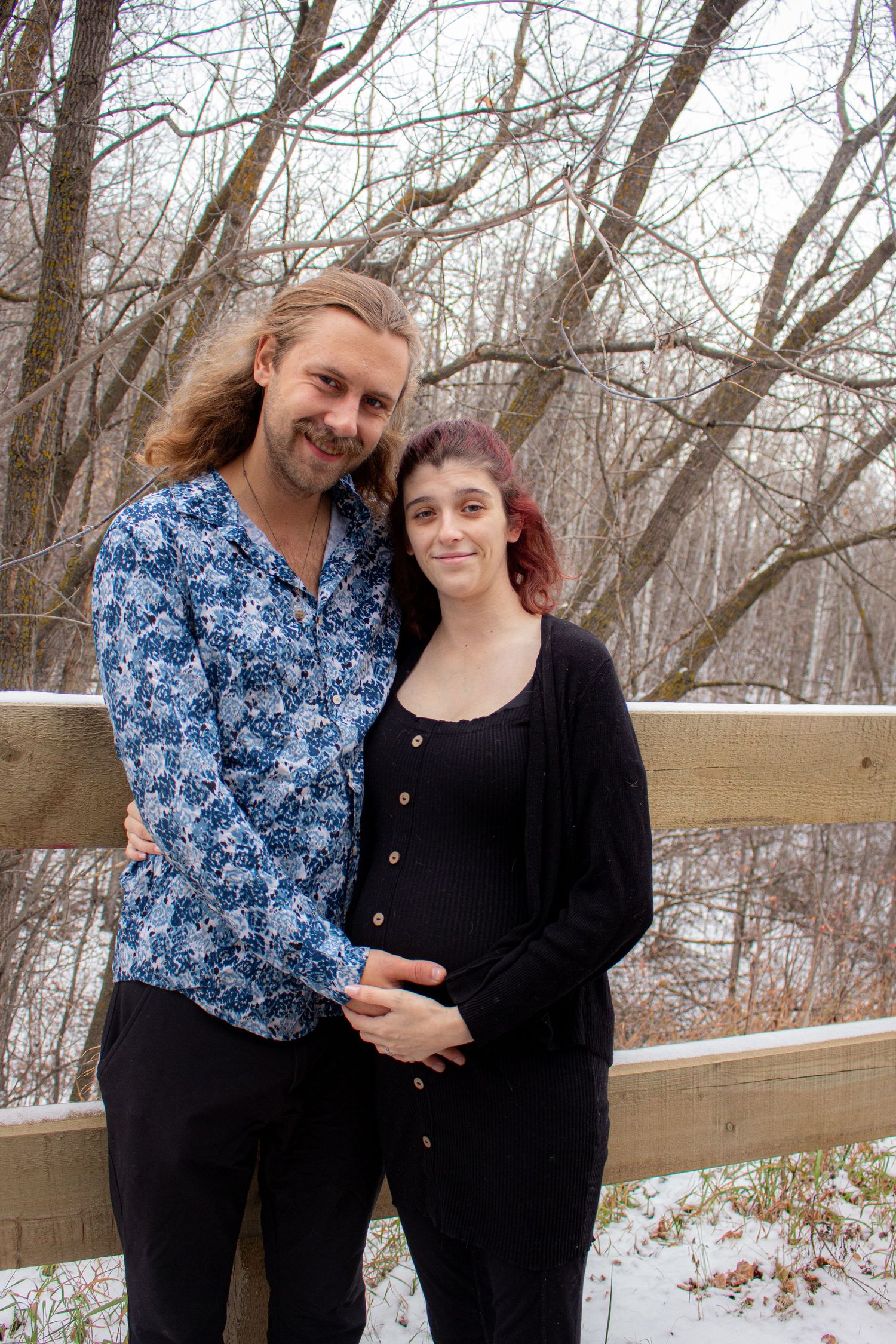 A couple poses for a winter maternity photo against a snowy woodland backdrop with wooden fence.
