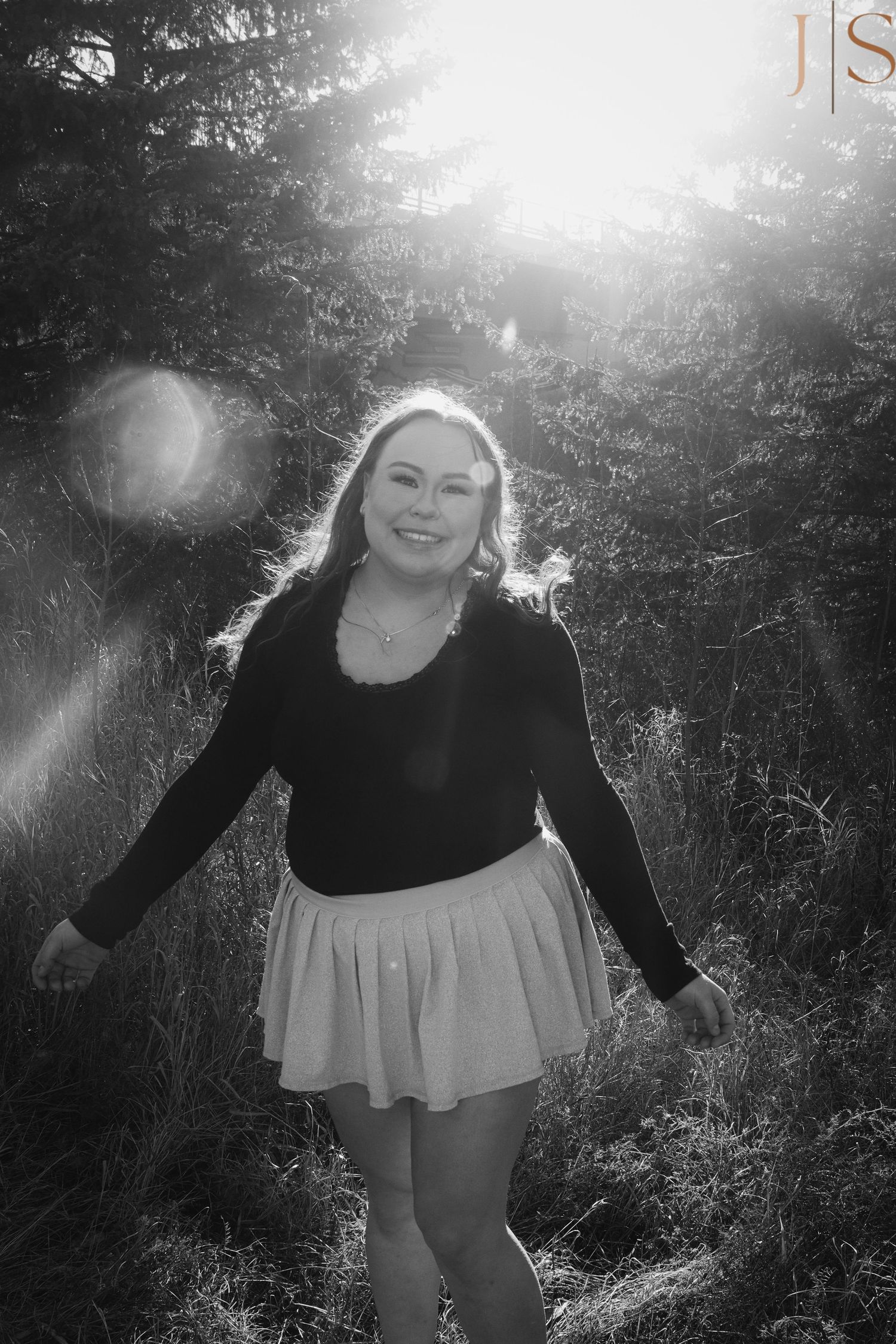 A black and white photo of someone in a dark top and light skirt dancing in sunlit woods with lens flare effects.