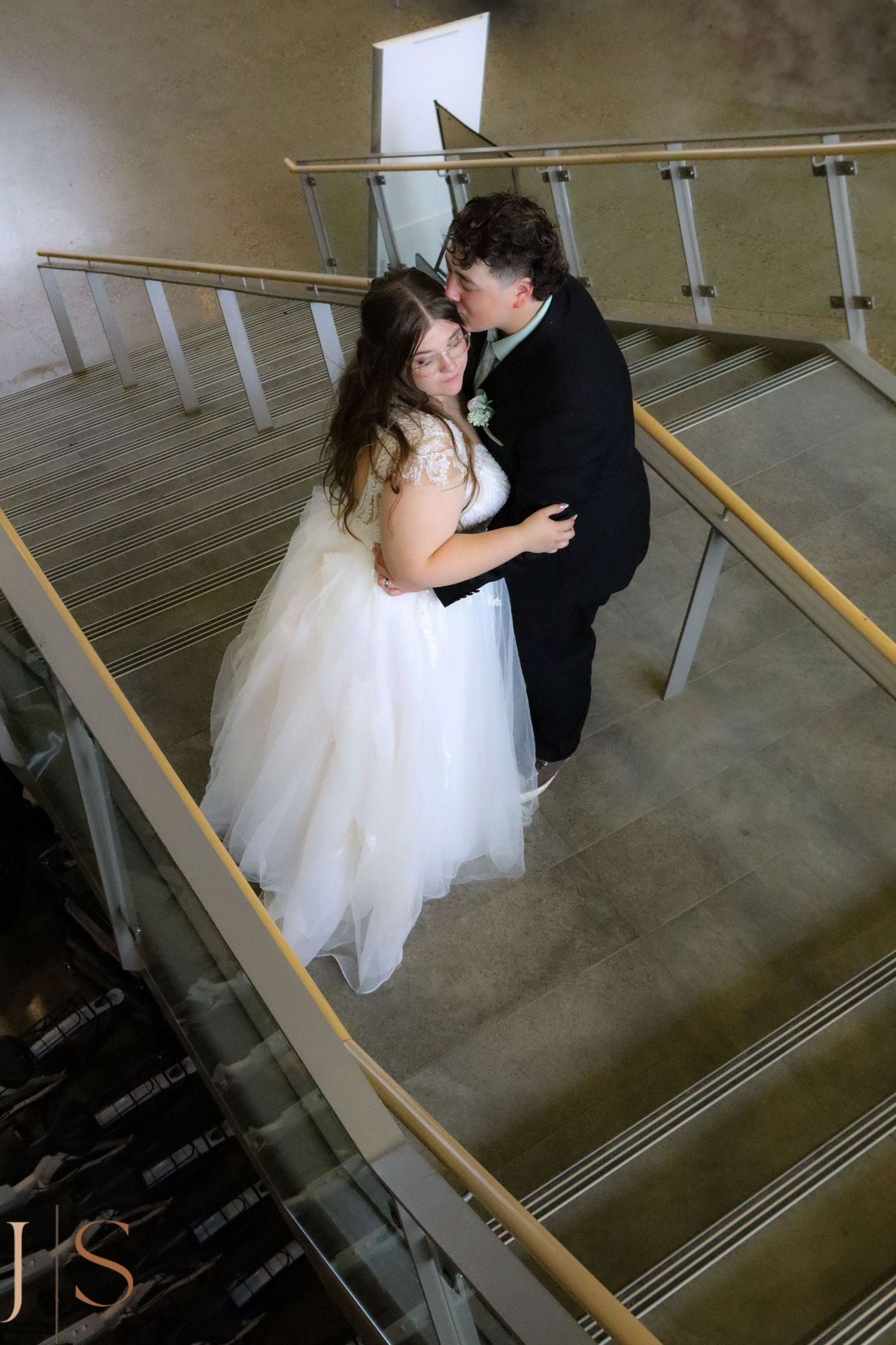 A bride in a white gown and groom in black suit share an intimate moment on a staircase with metal railings.