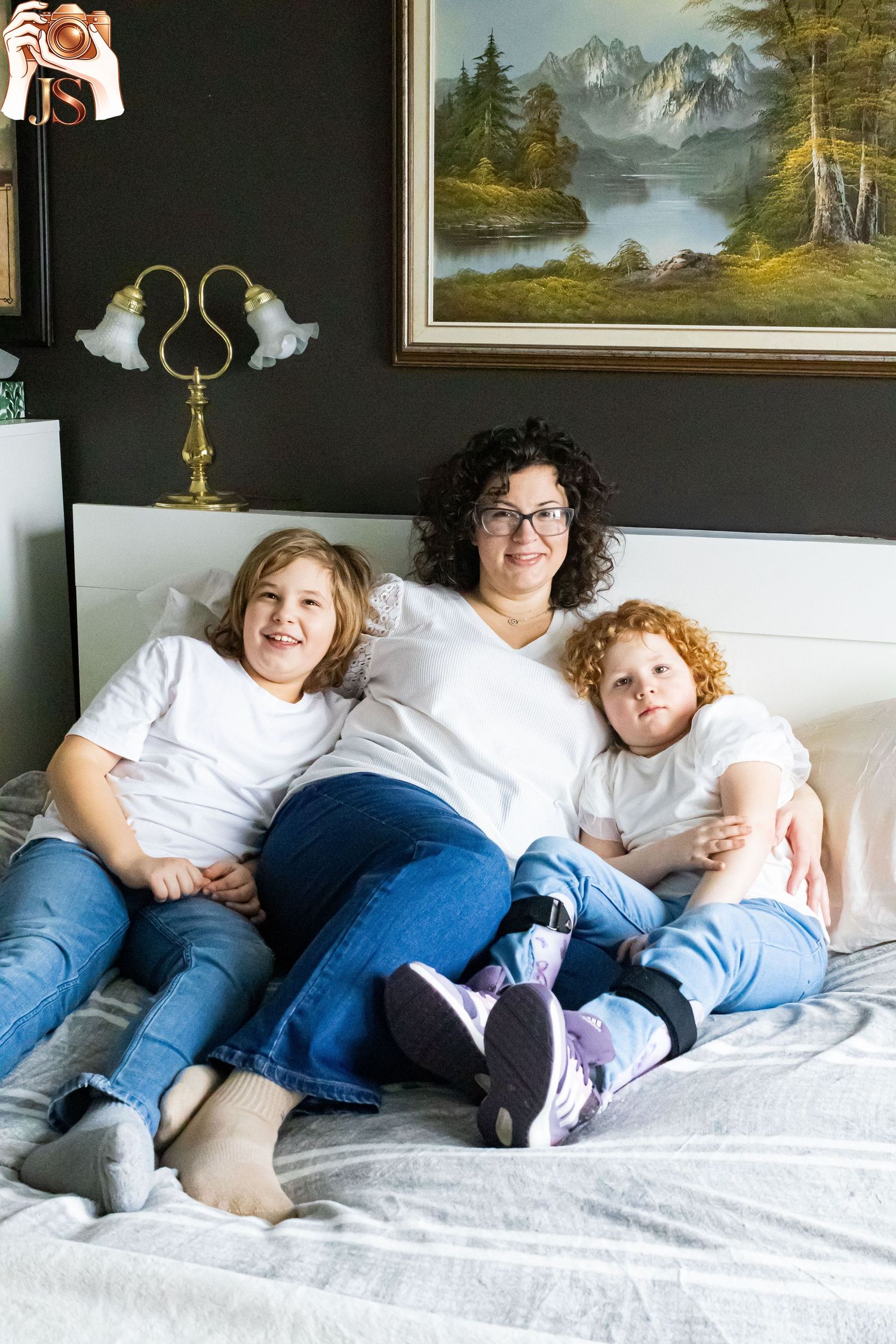 A cozy family photo session shows people lounging together on a white bed with landscape paintings on the dark wall.