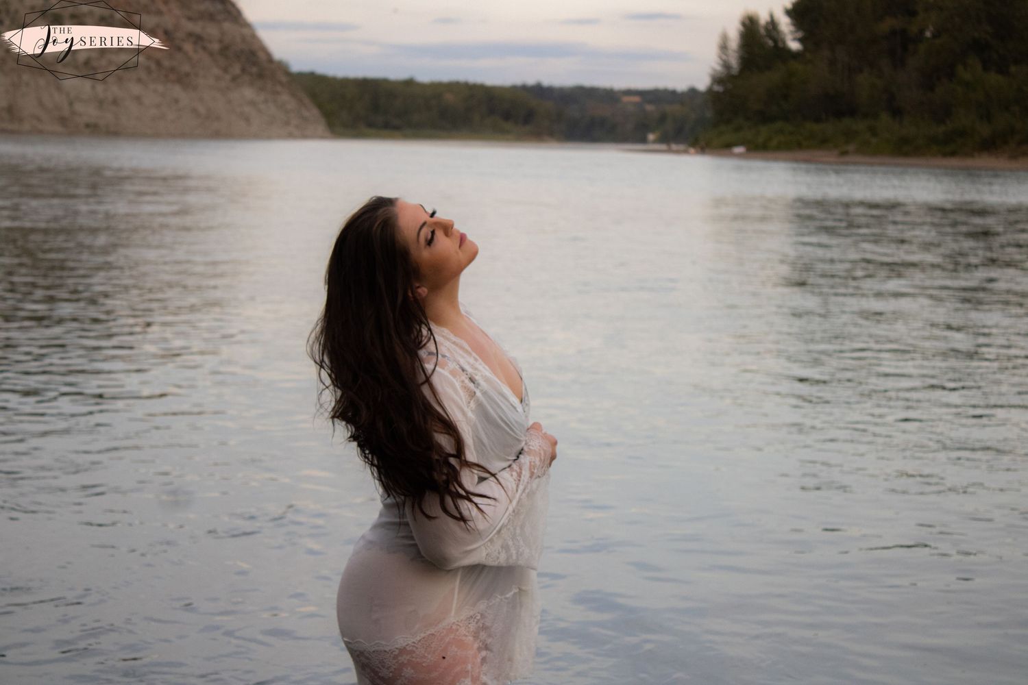 Someone in a white dress poses at the edge of a lake with mountains in the background during sunset.