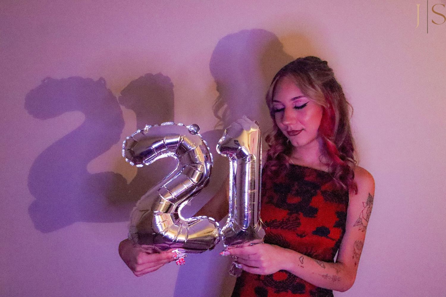 A person in a red and black dress holds silver number 21 balloons at a celebration with purple lighting.