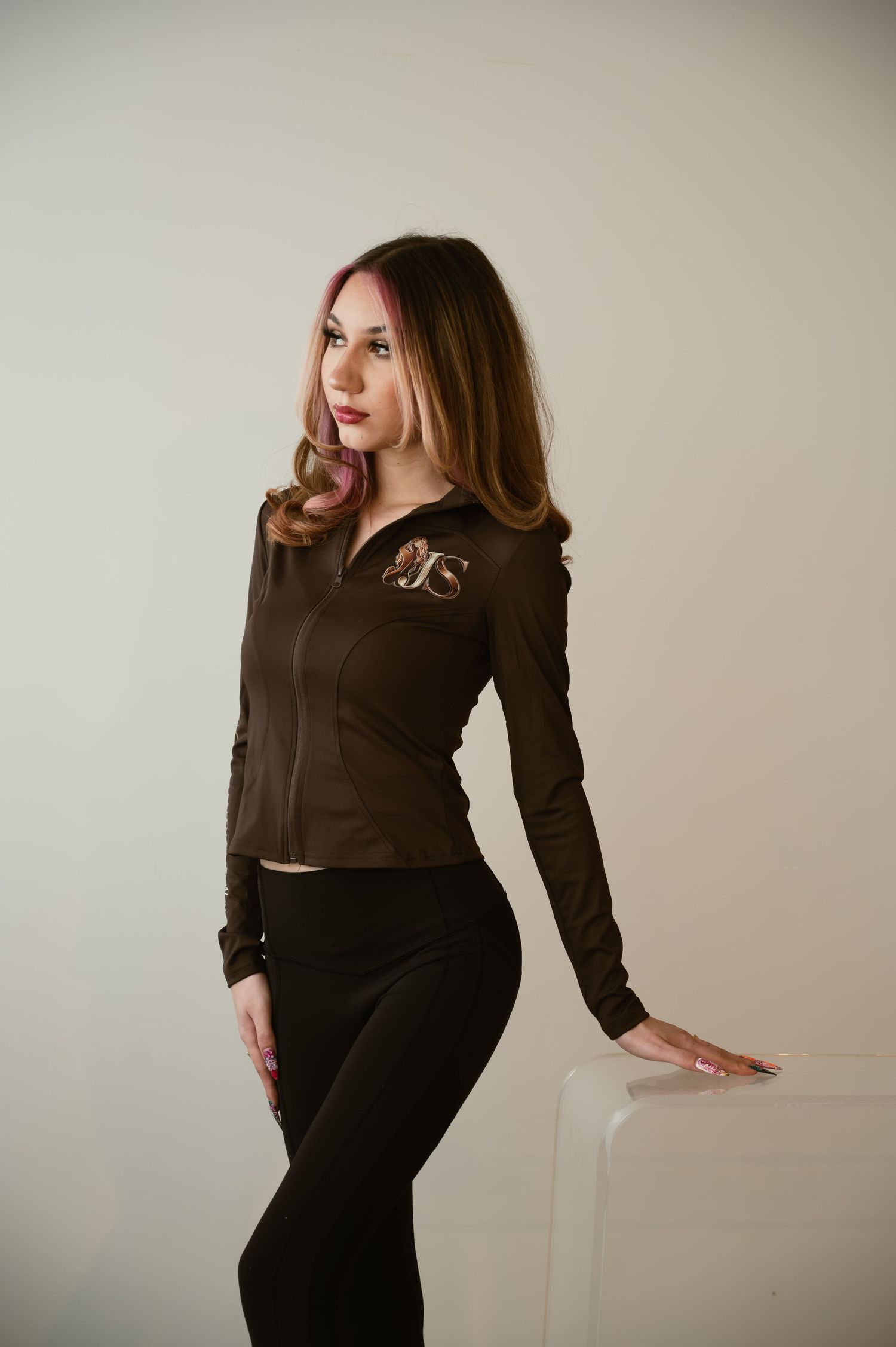 Young woman in brown zip-up jacket and black leggings poses confidently in a bright studio setting.