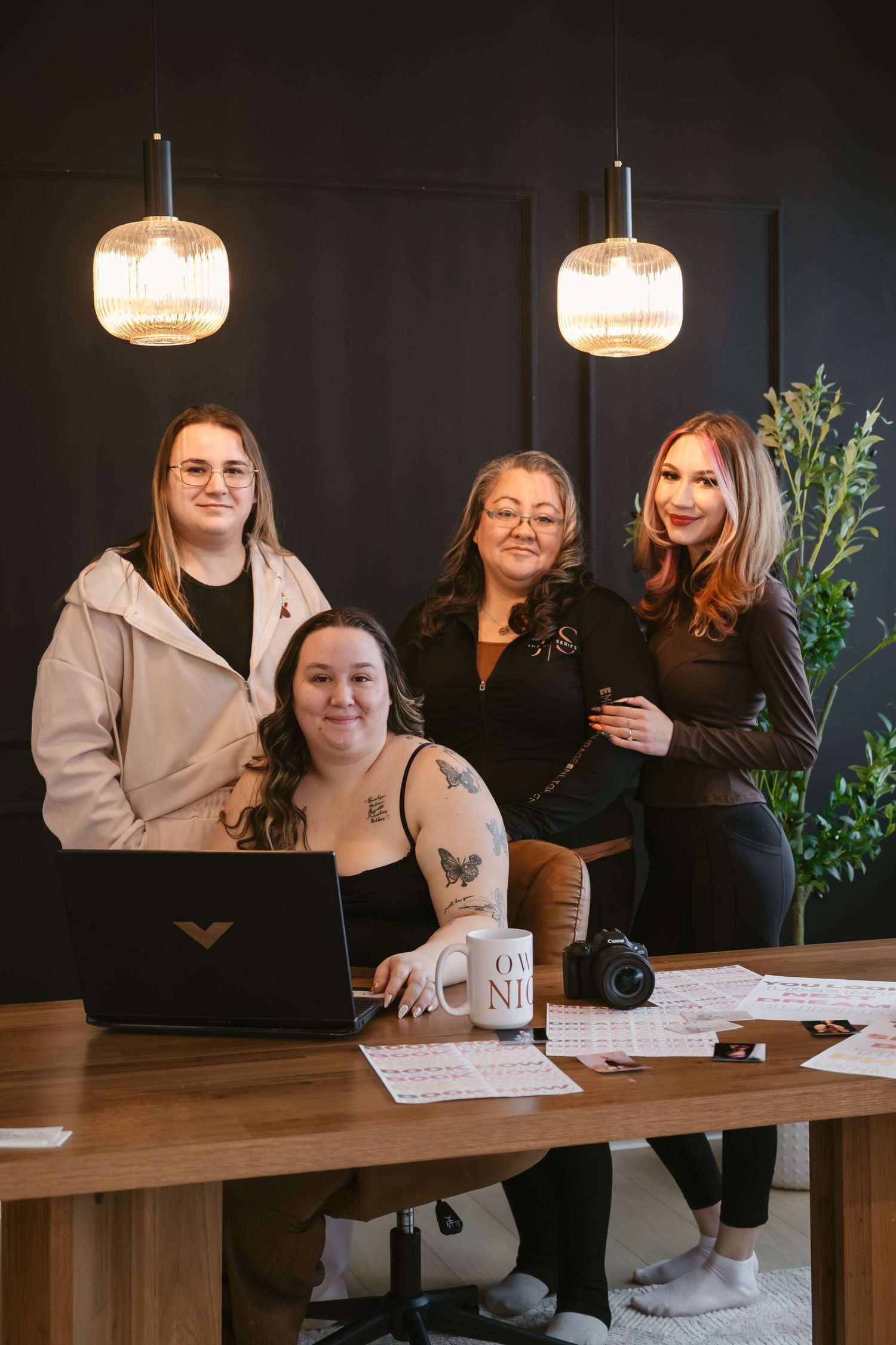 Four women pose together at a wooden desk with a laptop, camera, and papers, under warm pendant lights in a modern dark office.