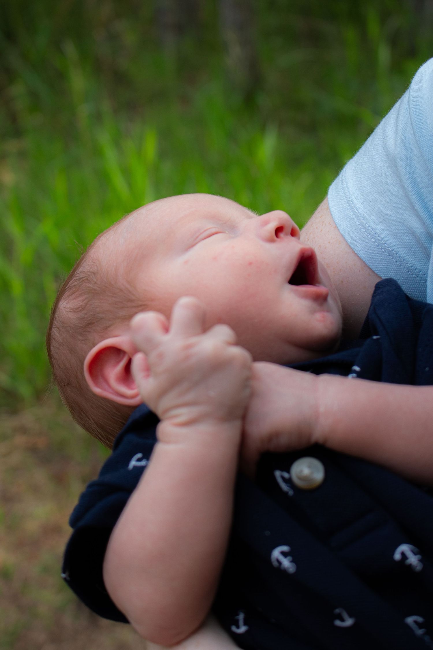 A baby in navy anchor print clothing nurses outdoors against green foliage.