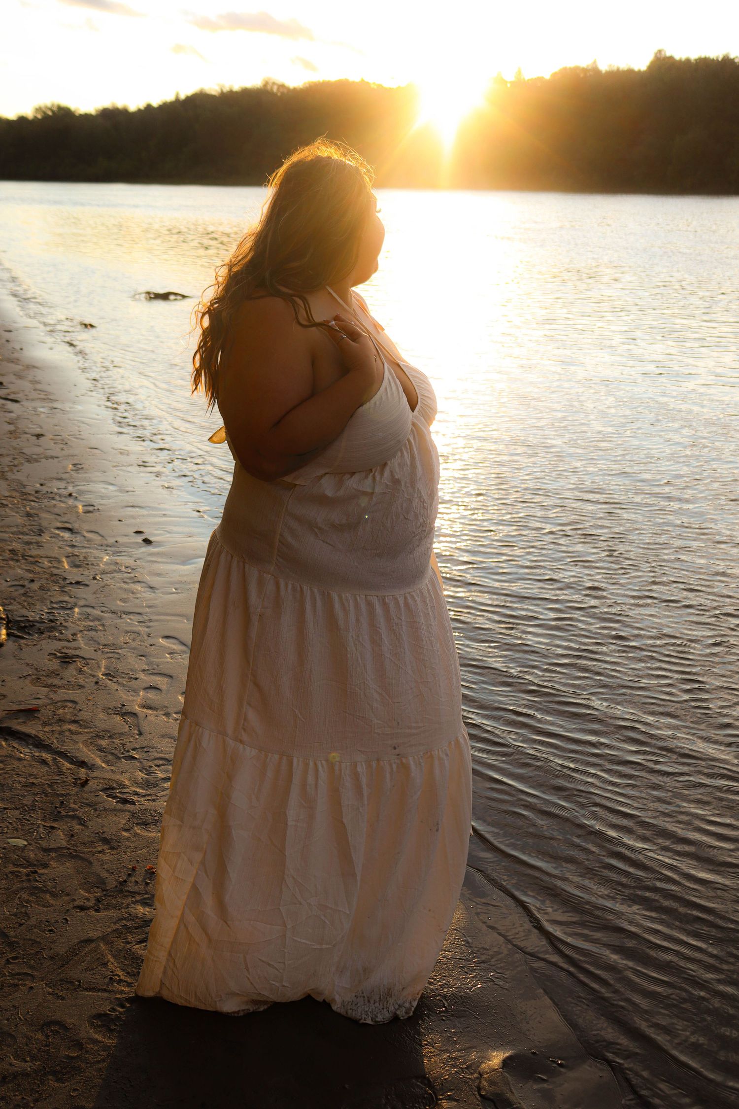 A silhouetted figure in a flowing white dress stands at the water's edge during sunset, creating a peaceful lakeside scene.