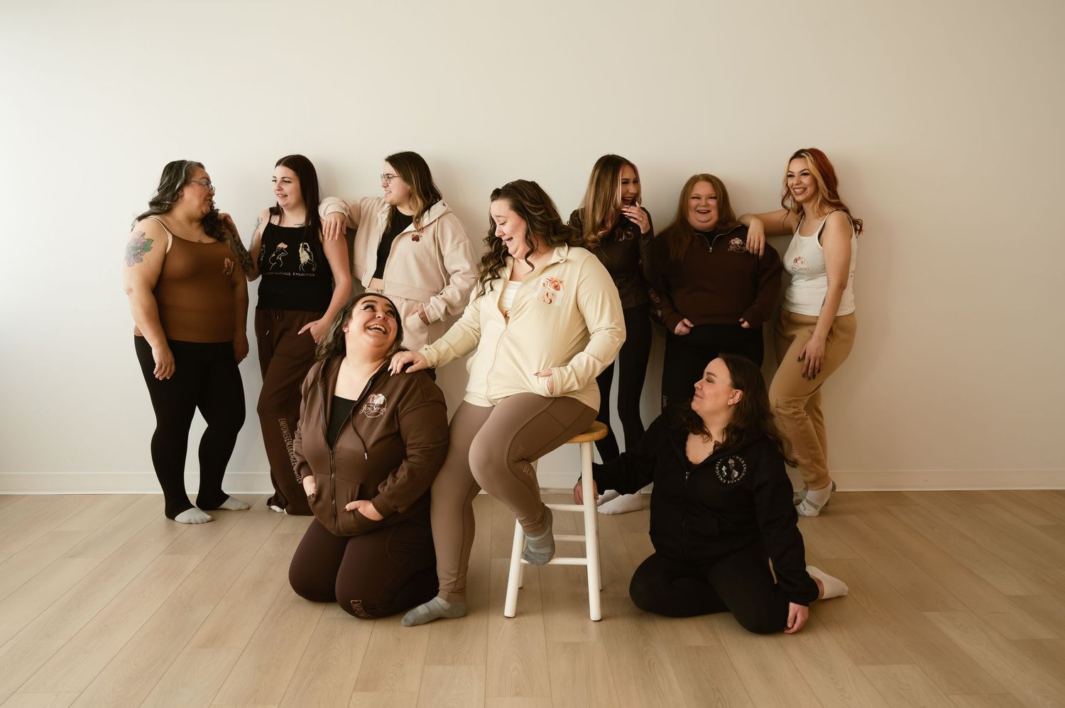Group of women in neutral brown tones posing together in a bright studio, some seated and some standing, laughing joyfully.