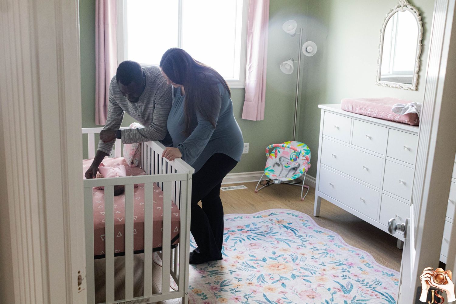 A parent leans over a white crib in a nursery room decorated with a floral rug and white dresser.