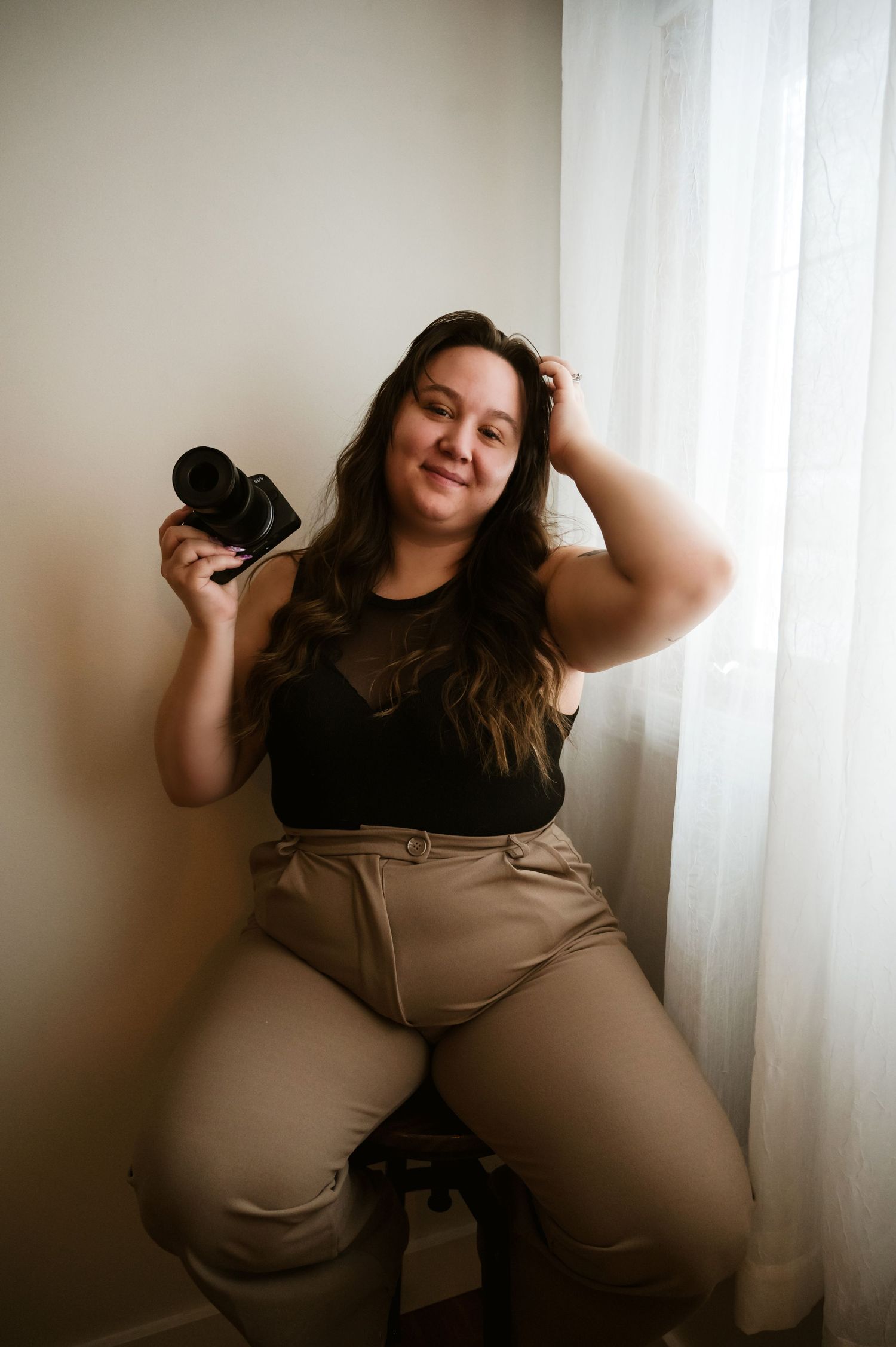 A photographer in a black top and beige pants sits casually by a window holding a camera.