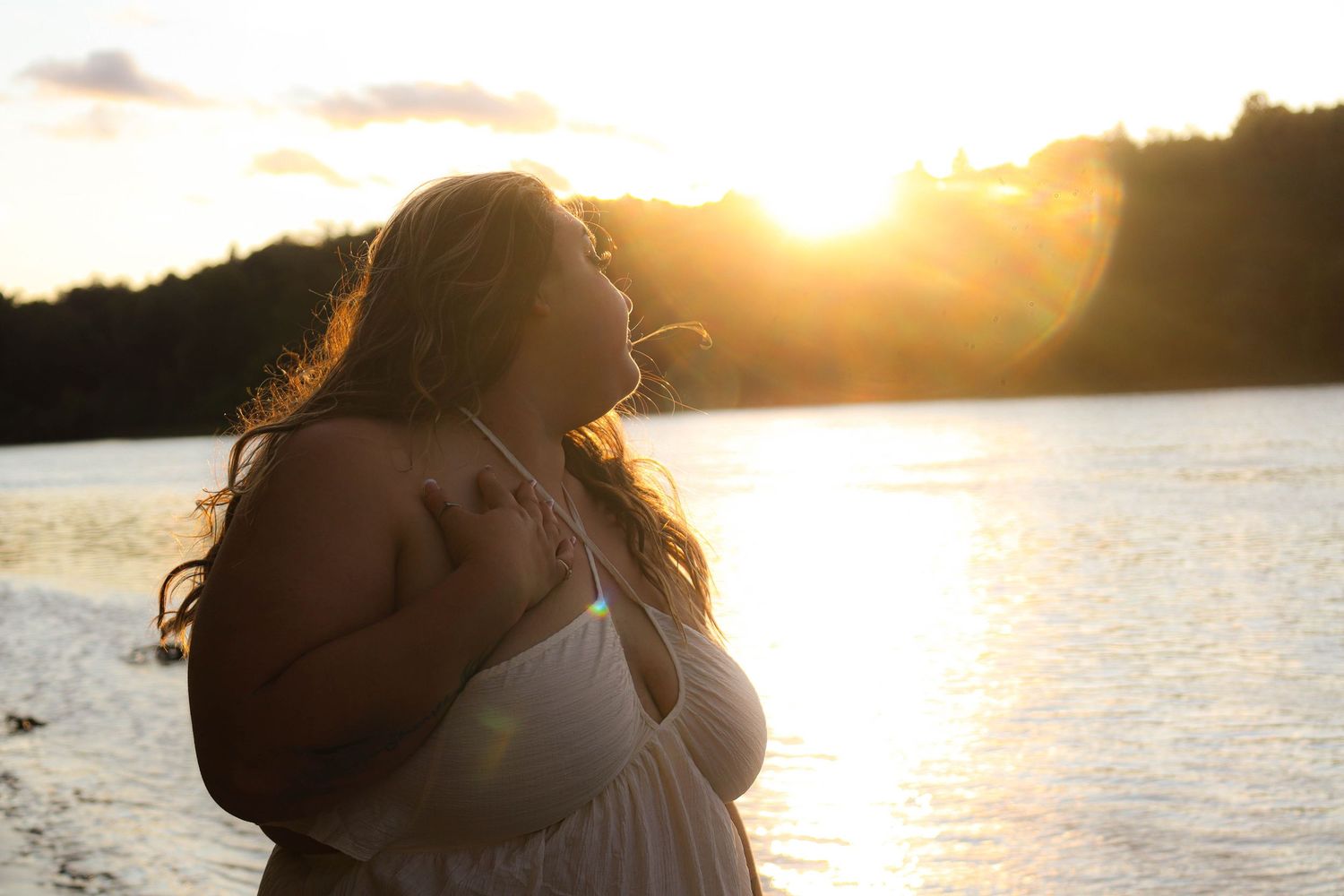 Silhouetted figure in white dress stands by lake at sunset with sun rays creating golden rim lighting.