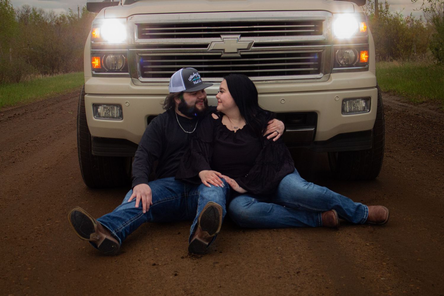 A couple sits on a dirt road in front of a white Chevrolet truck with headlights on.