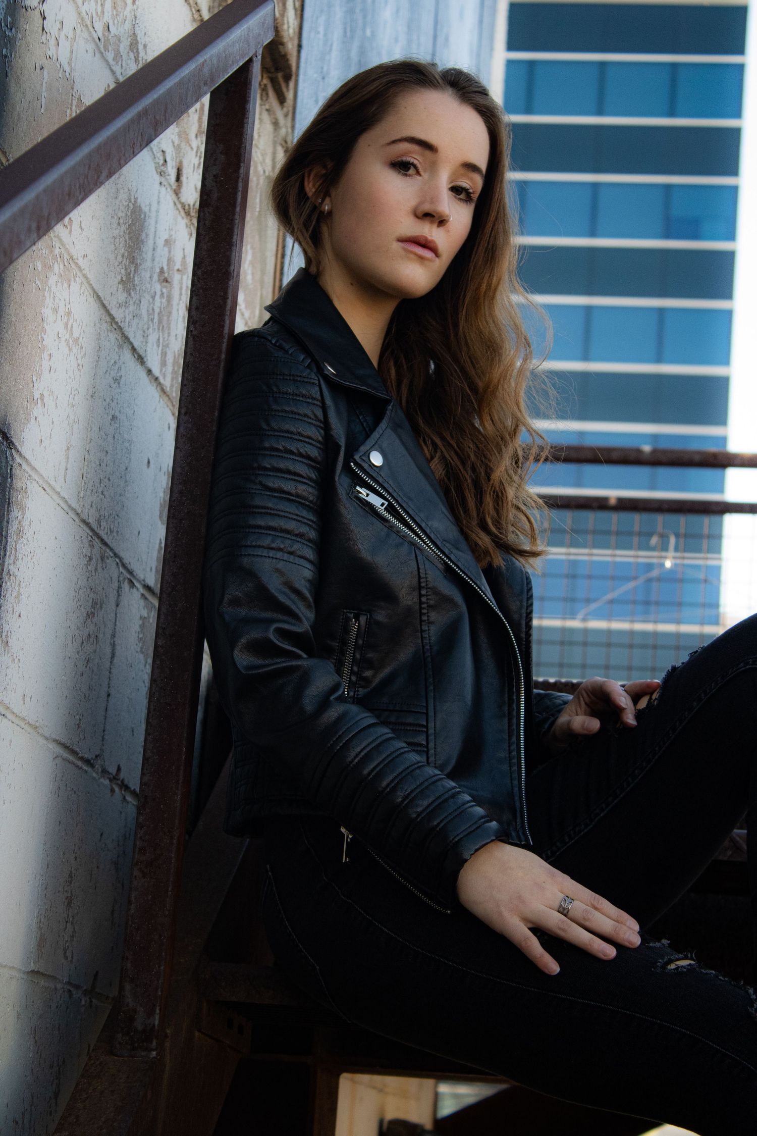 A fashion model in a black leather jacket poses against an industrial backdrop with metal stairs.