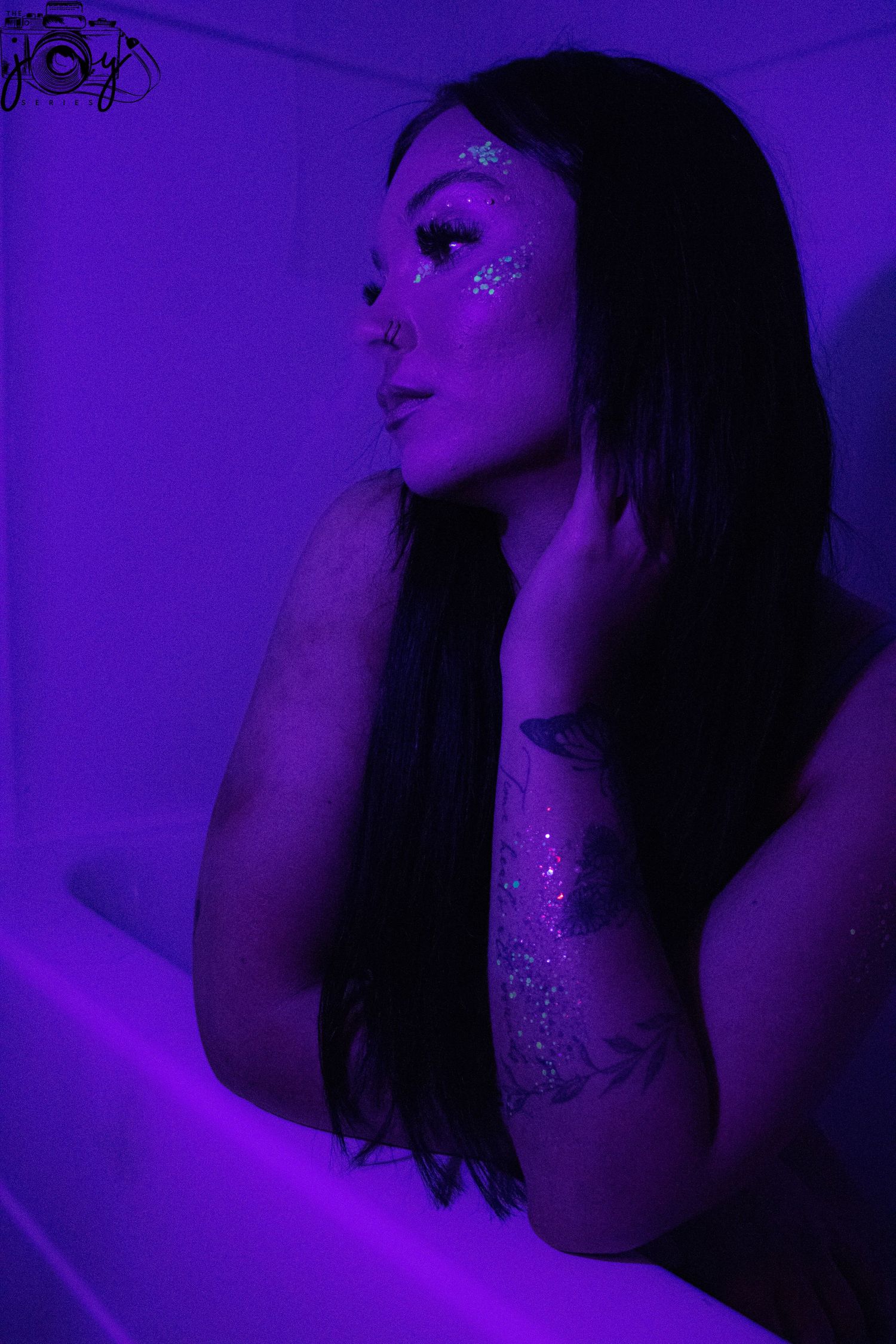 Artistic portrait in purple lighting shows a person with long dark hair and glitter makeup posing dreamily.