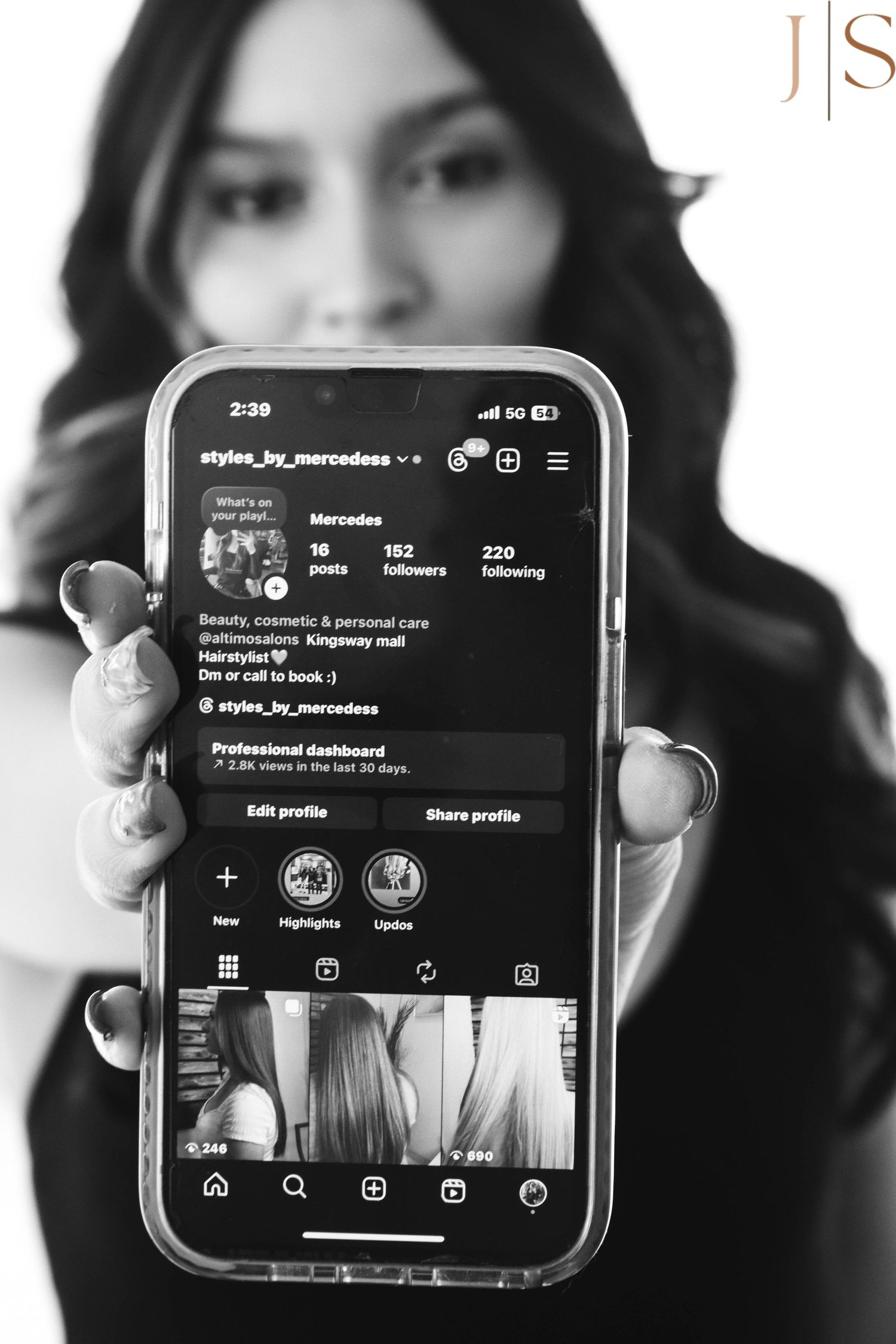 A person's hand holding a smartphone displaying a social media profile interface in black and white monochrome.