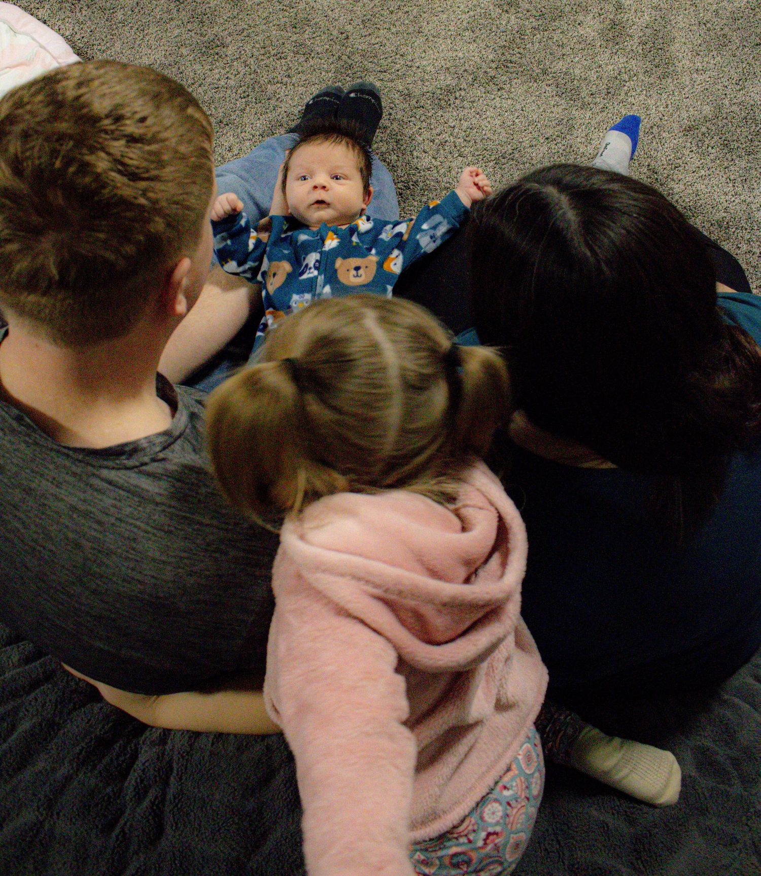 Family members sitting together on carpet looking down at baby in blue pajamas.