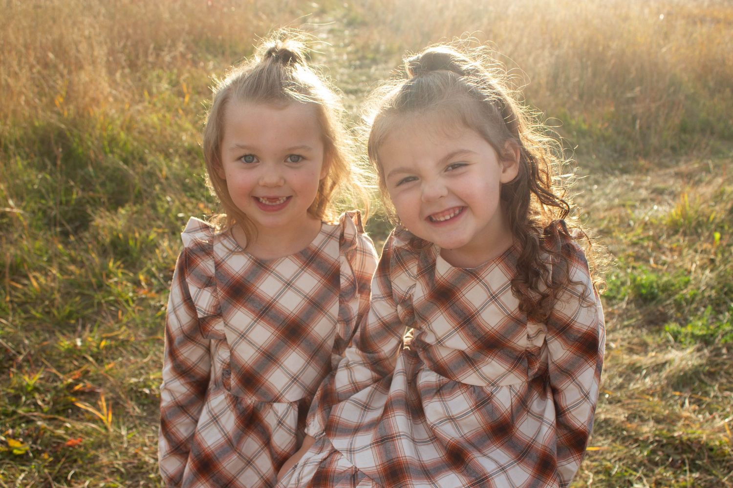 Two children wearing matching plaid dresses pose outdoors in golden sunlight.