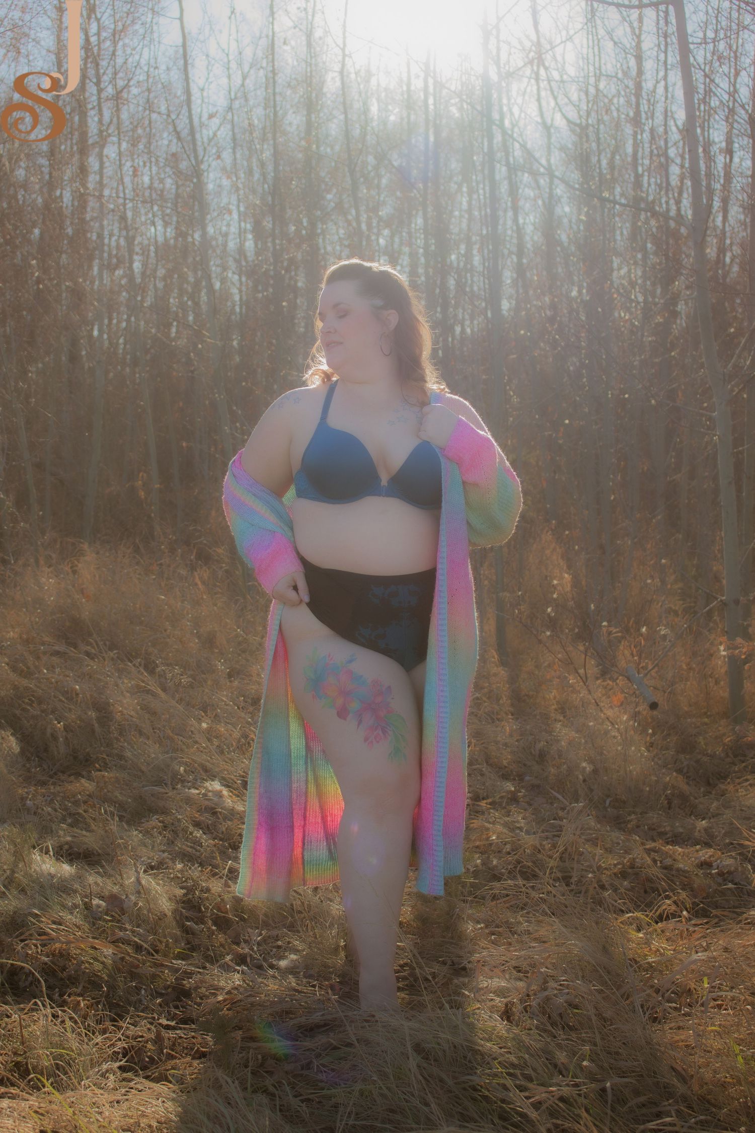 A plus-size model in black swimwear and colorful tie-dye cover-up poses in a misty forest clearing at sunset.