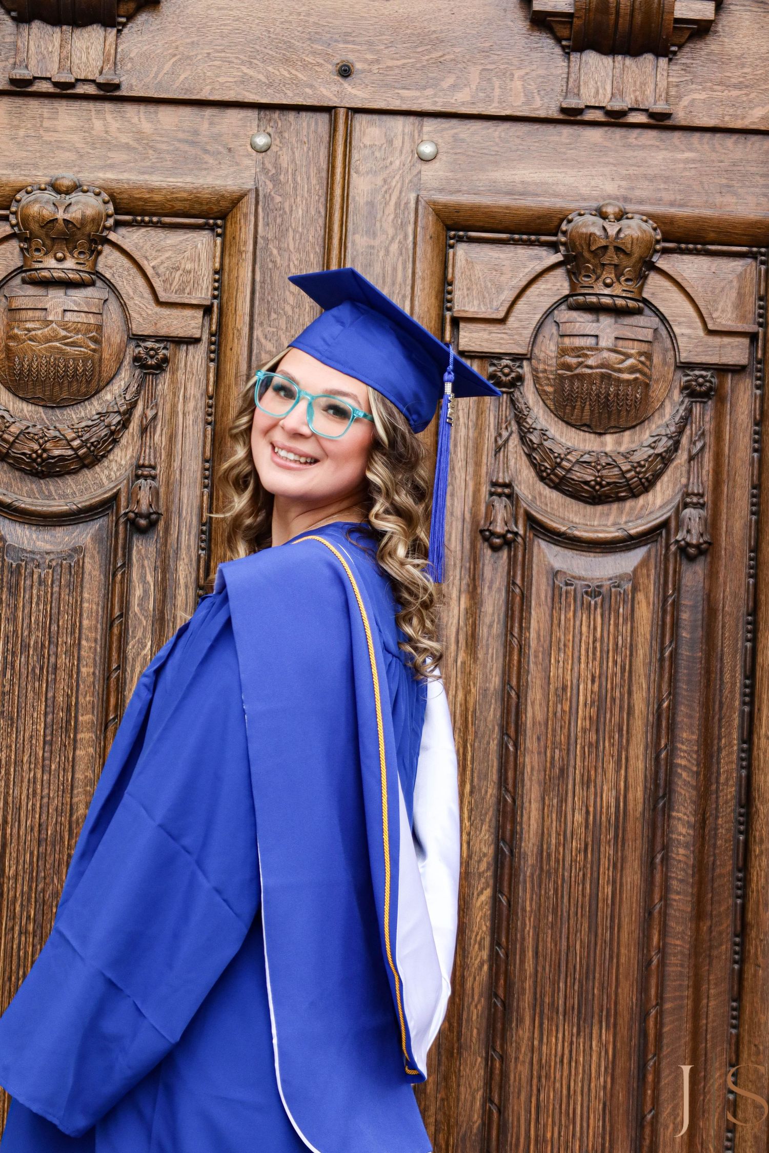 A graduate poses in front of ornate wooden doors wearing traditional blue graduation regalia.
