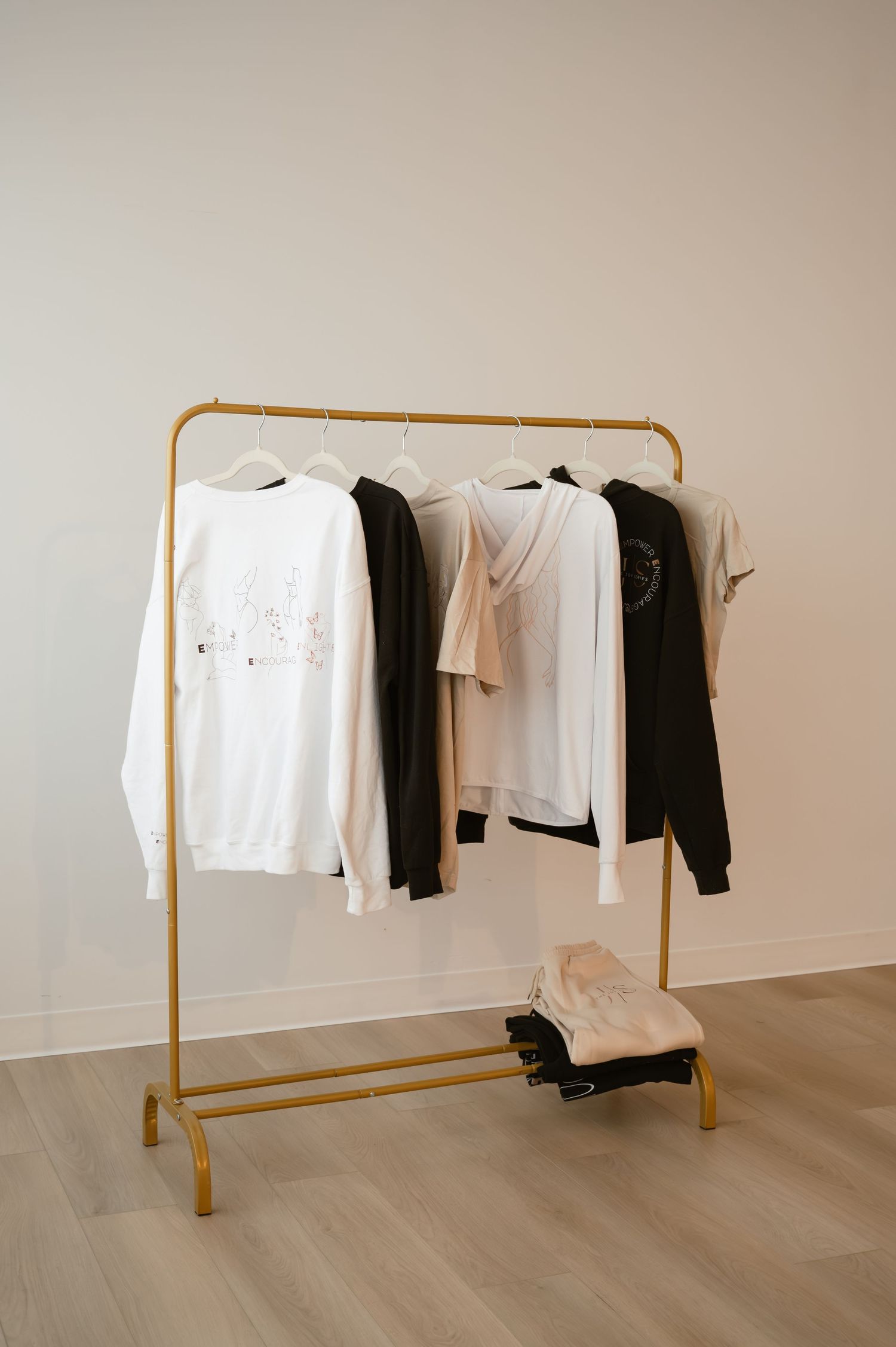 Gold clothing rack with white, black, and beige sweatshirts and tops displayed against a neutral wall.