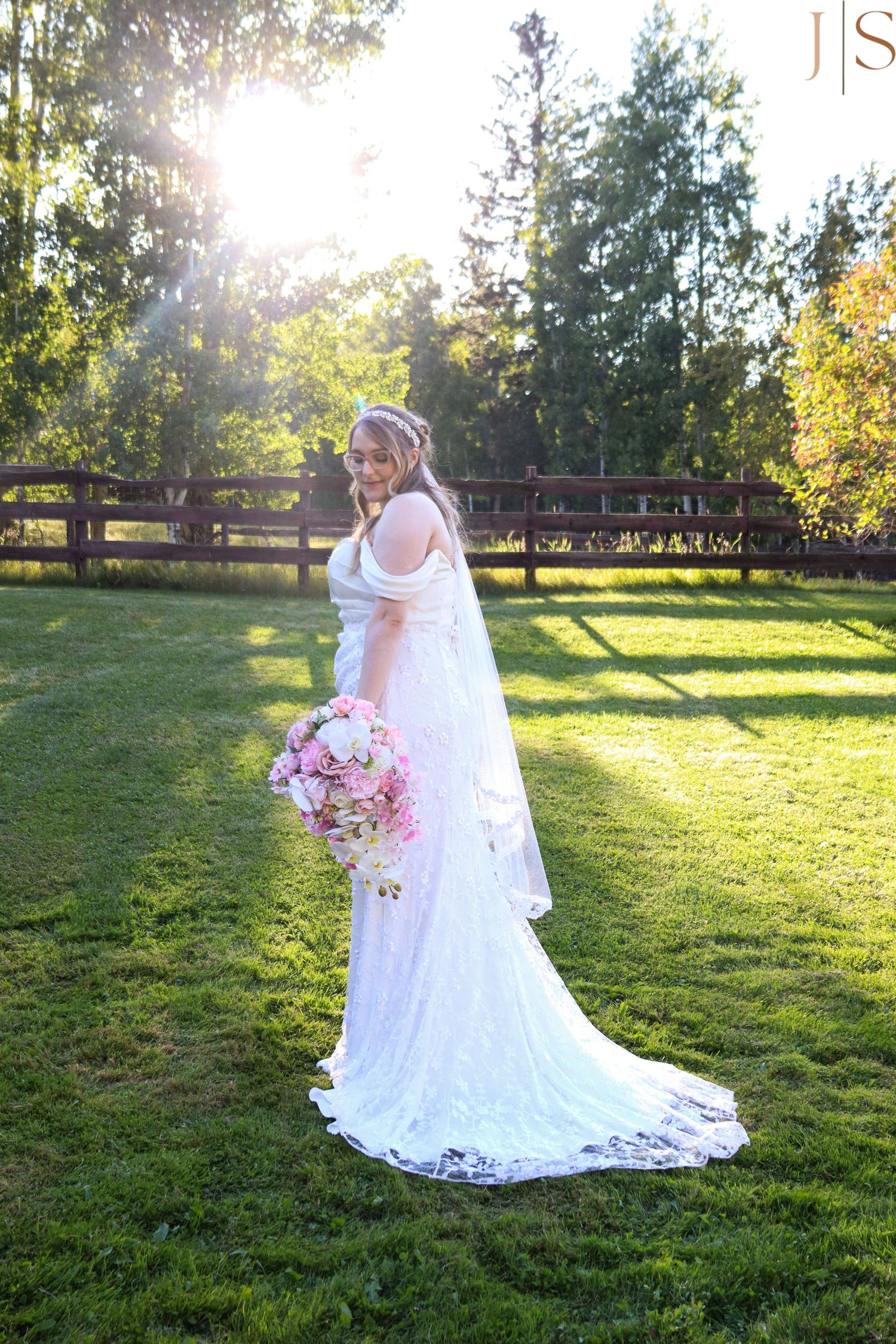 A bride in a flowing white lace gown poses in a sunlit garden setting with a pink and white floral bouquet.