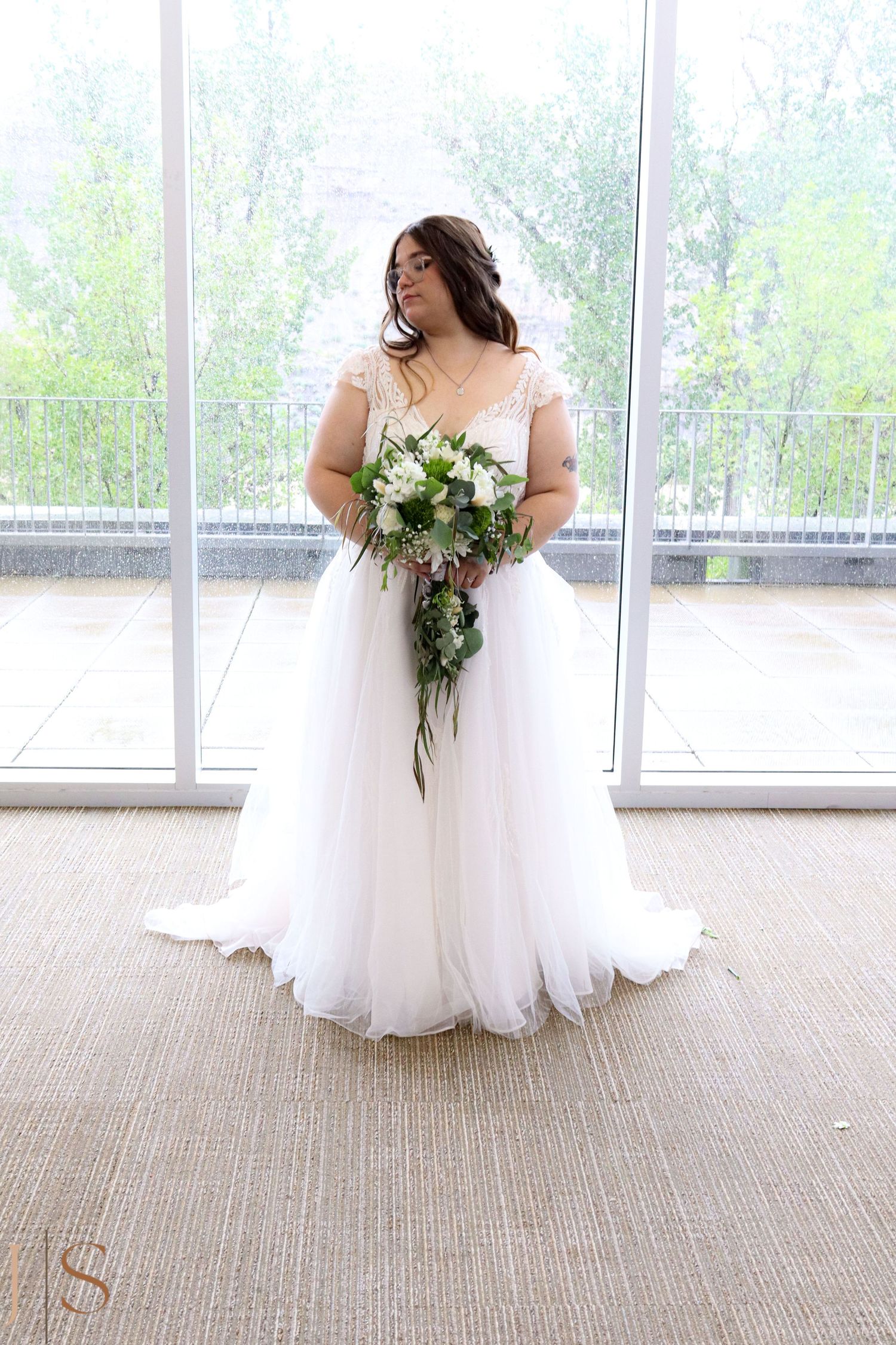 A bride in a flowing white gown holds a cascading bouquet while standing by large windows.