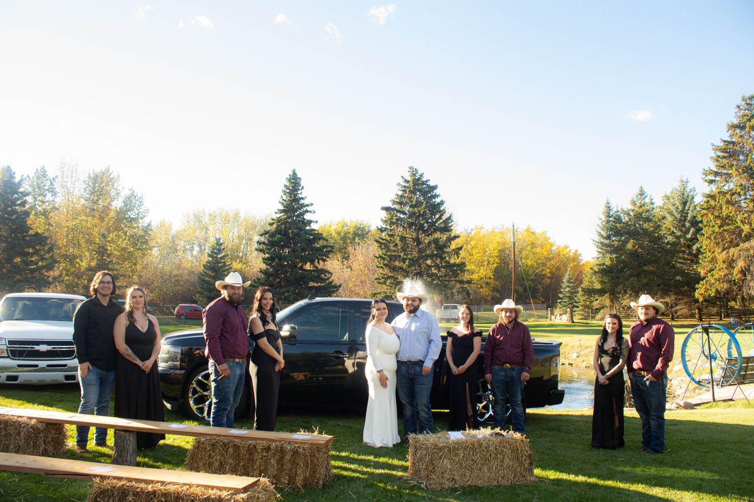 Outdoor country wedding scene with hay bales and guests gathering near parked cars.