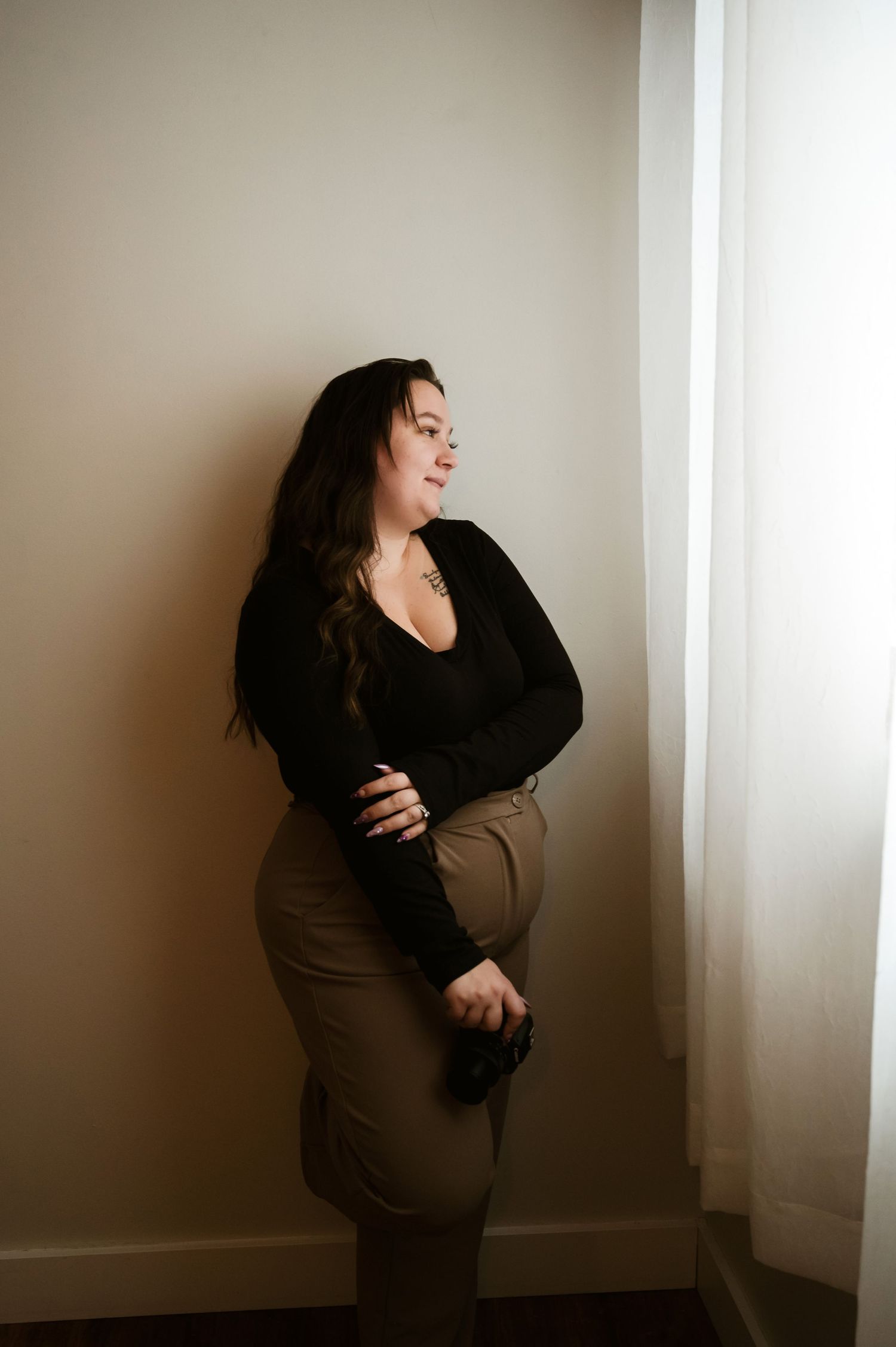 An artistic portrait with dramatic lighting shows a figure in a black sweater and brown skirt standing by a window.