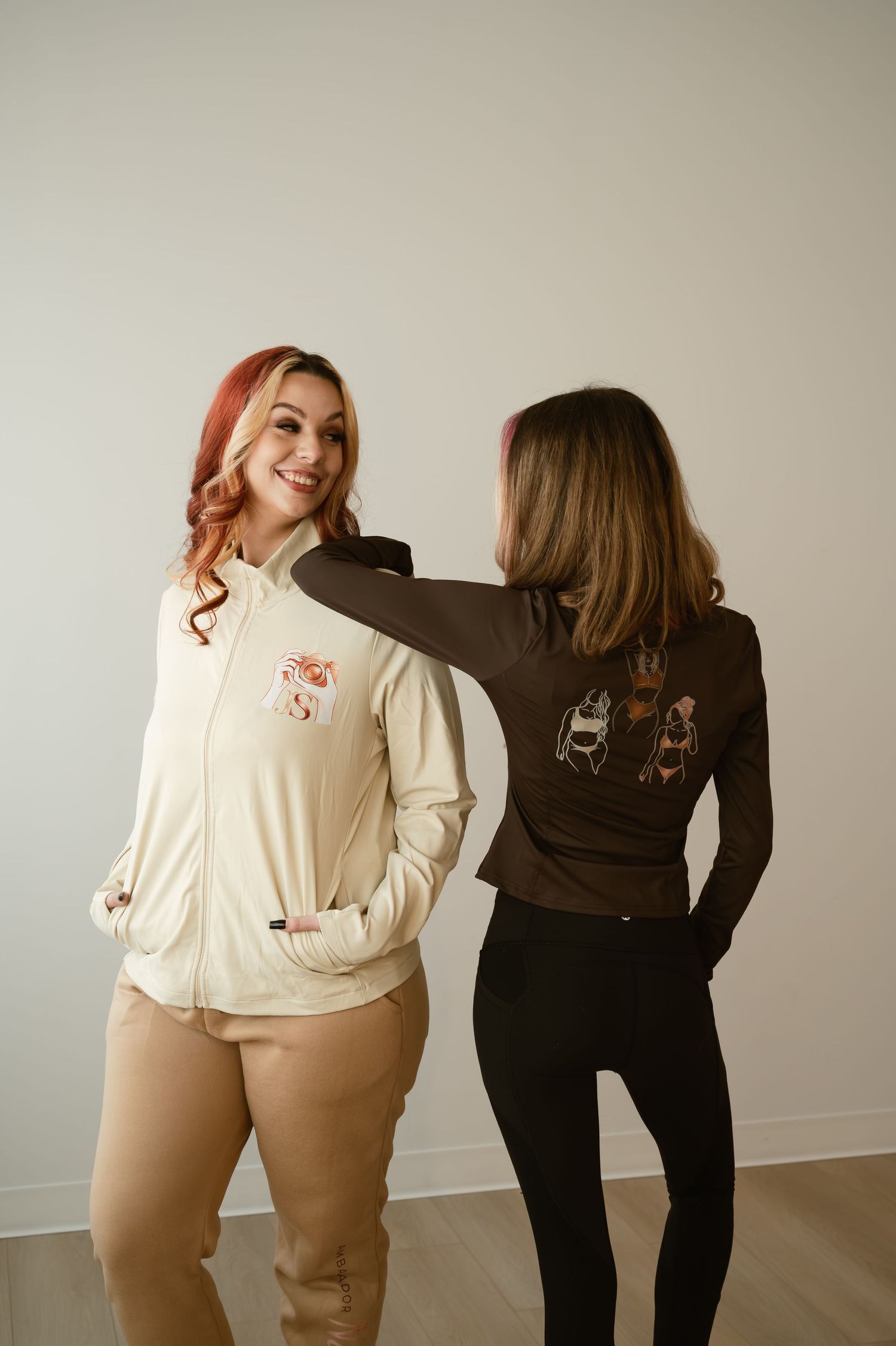 Two women modeling embroidered jackets, one cream zip-up and one brown long-sleeve, posing together against a white wall.