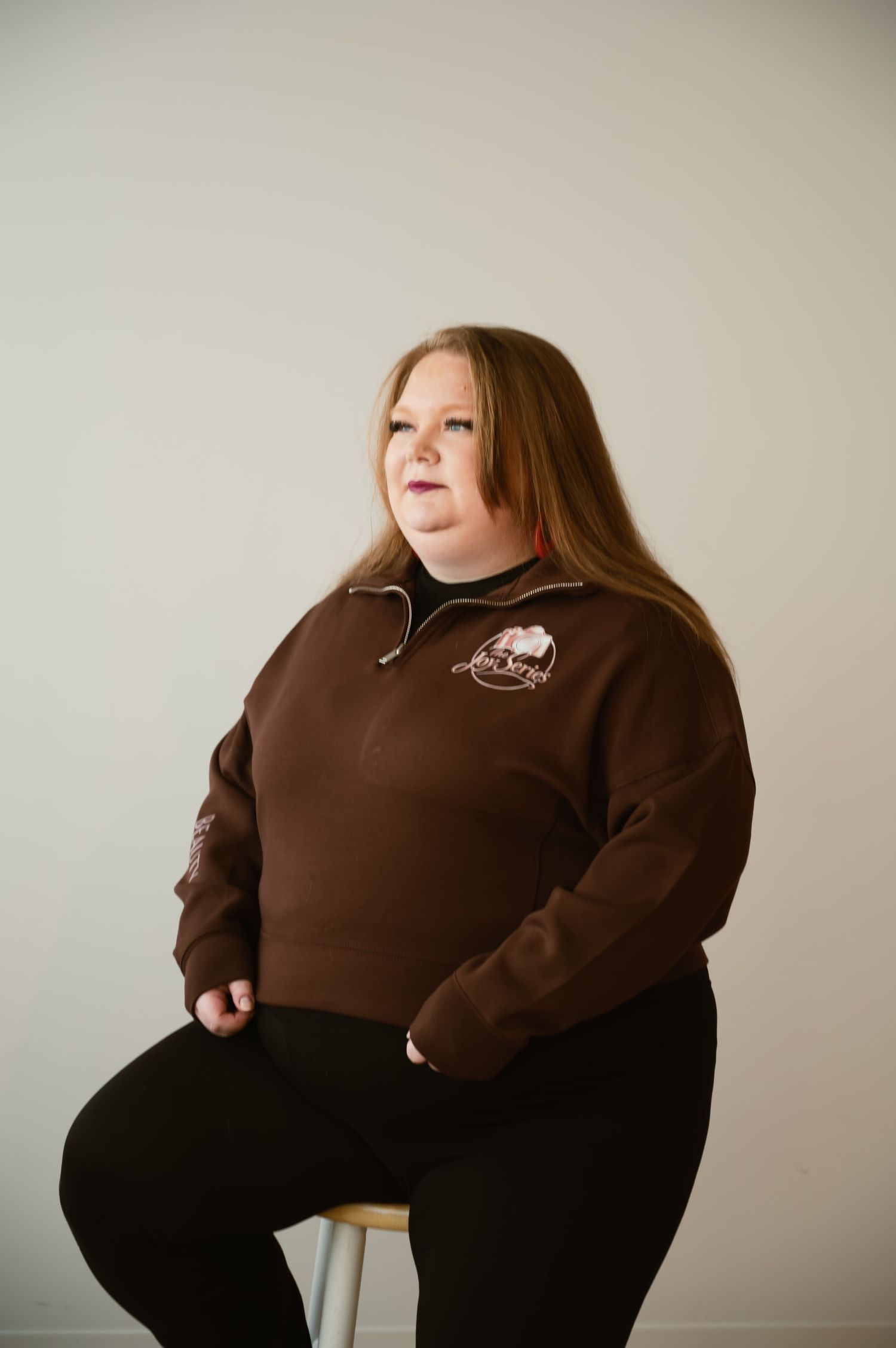 Woman in brown quarter-zip sweatshirt with embroidered logo seated on stool against neutral background.