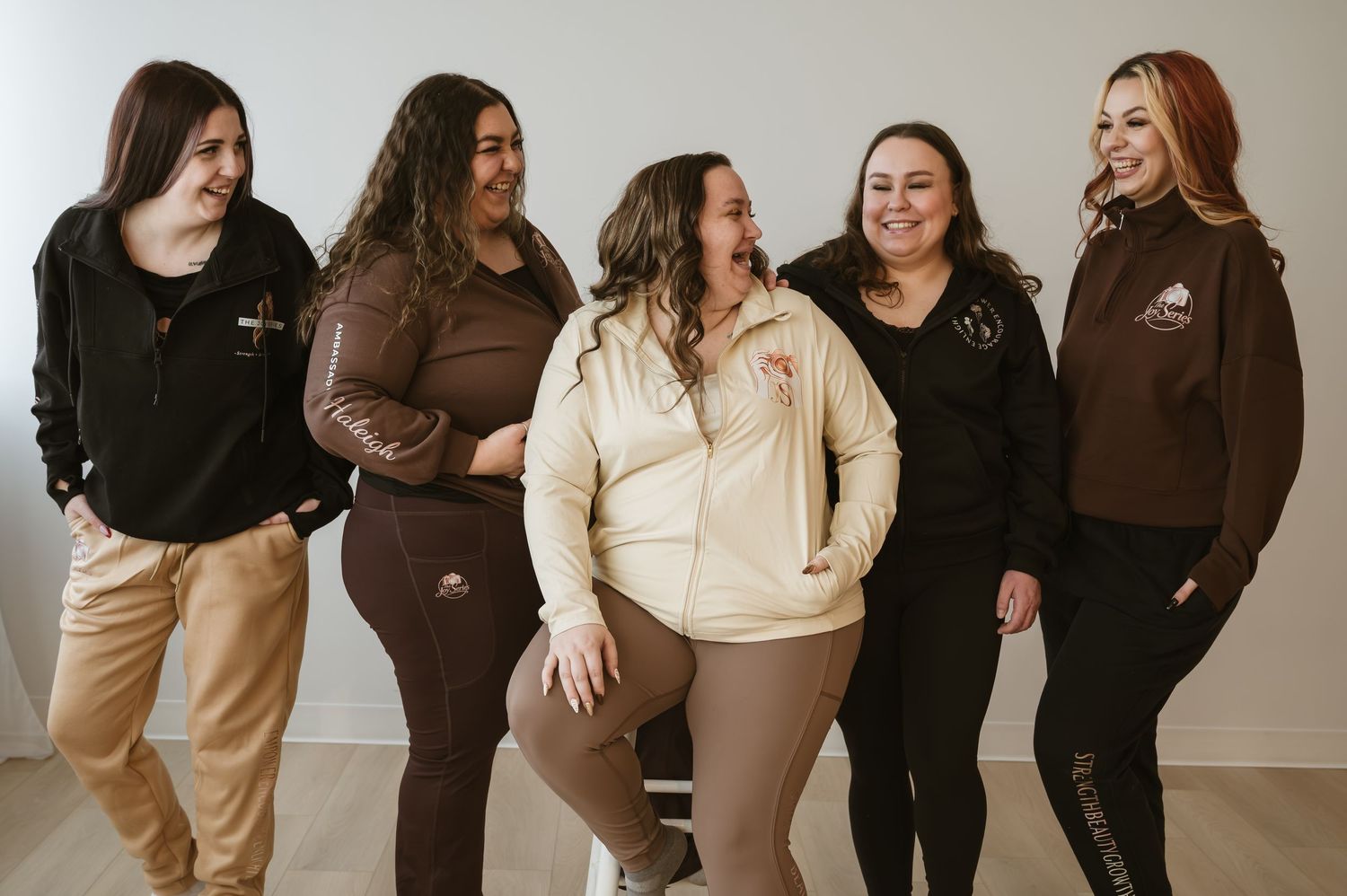 Five women in matching brown and cream branded apparel smile together in a bright studio setting.