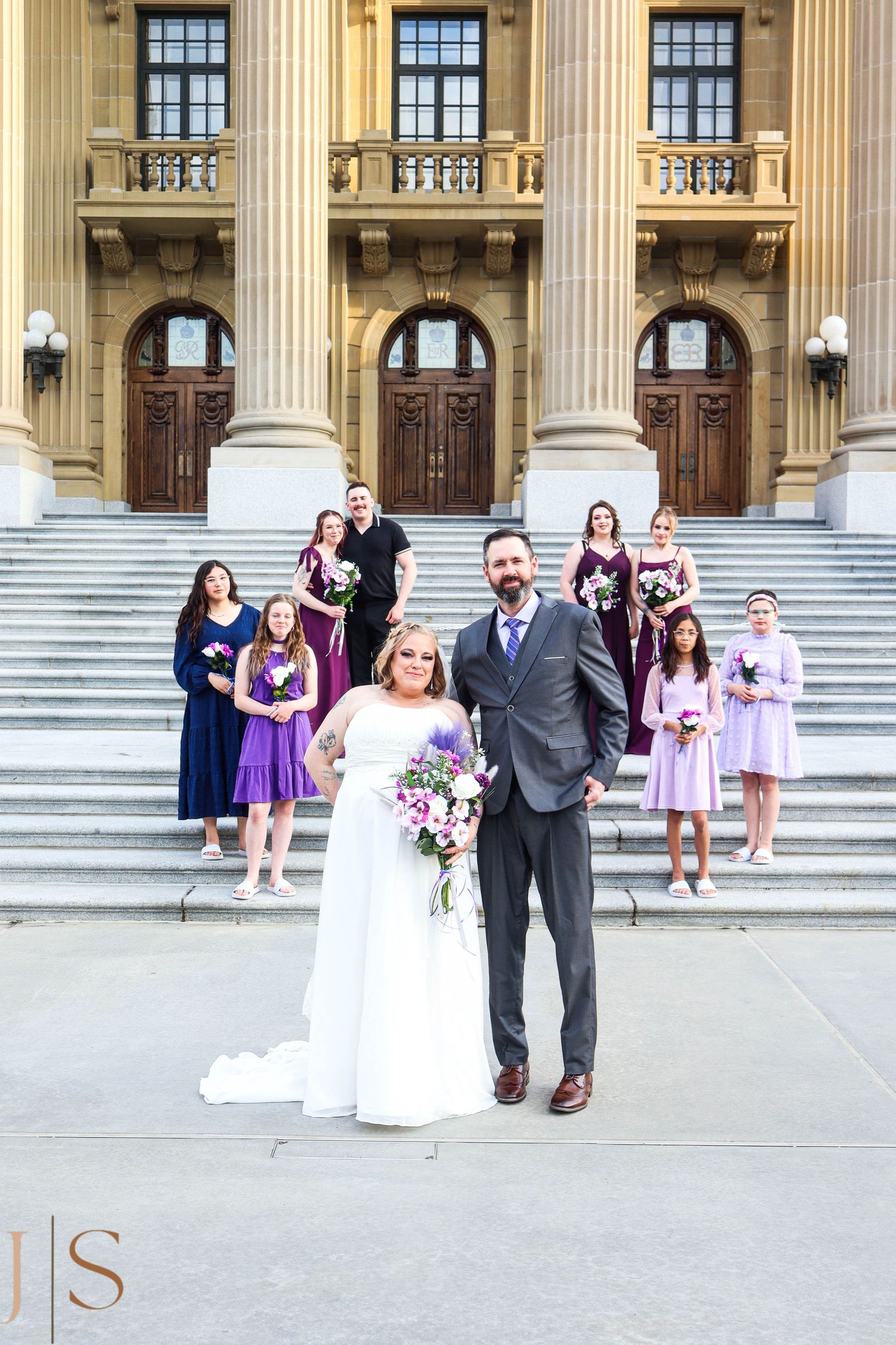 Wedding party gathered on grand stone steps of historic building with massive columns and elegant wooden doors.