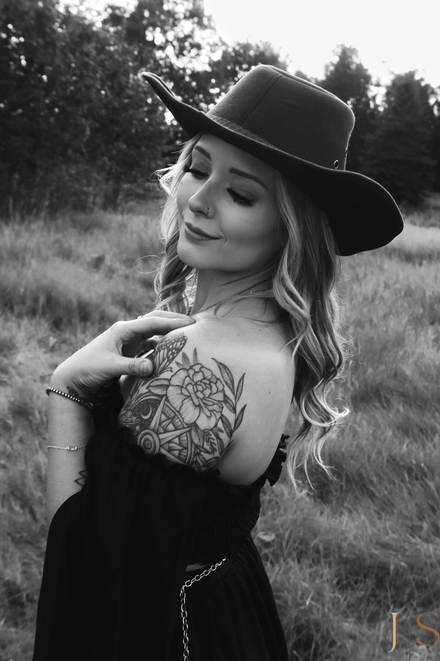 Black and white portrait with hat and visible shoulder tattoo in outdoor setting.