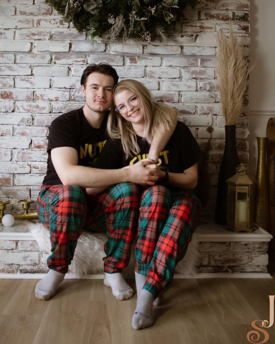 Couple wearing matching plaid pajama pants sits together against white brick wall with holiday decorations.