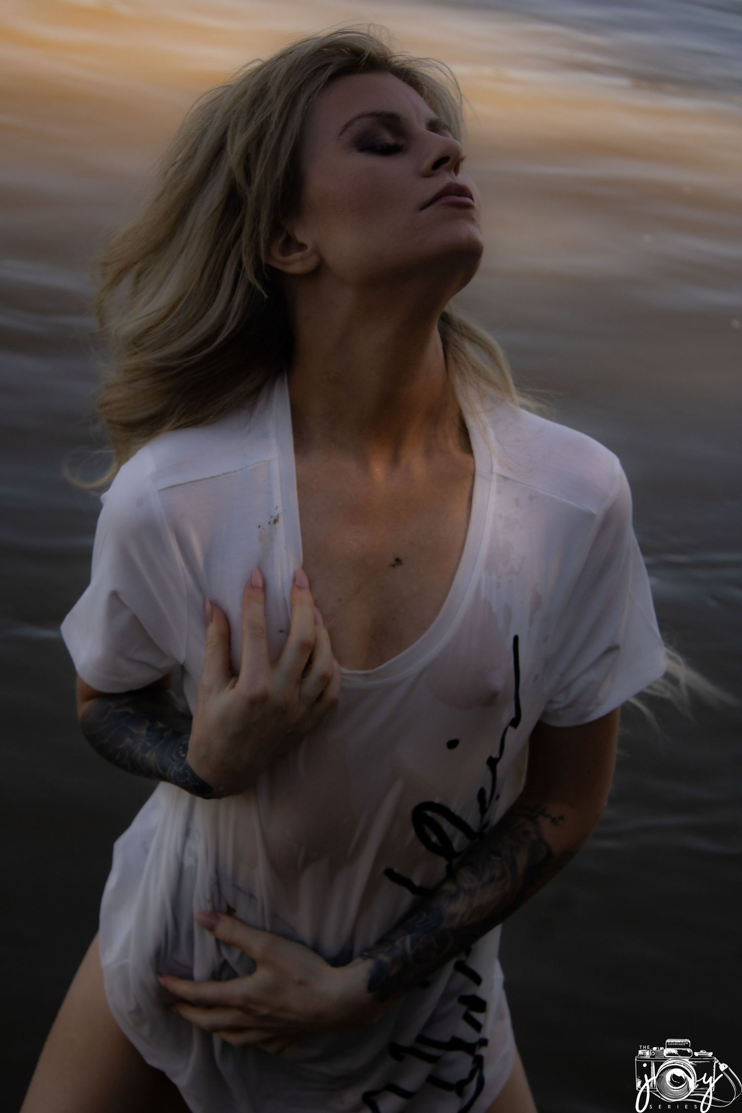 A person in a white shirt with tattoos poses dramatically against a moody sunset sky with eyes closed and windswept hair.