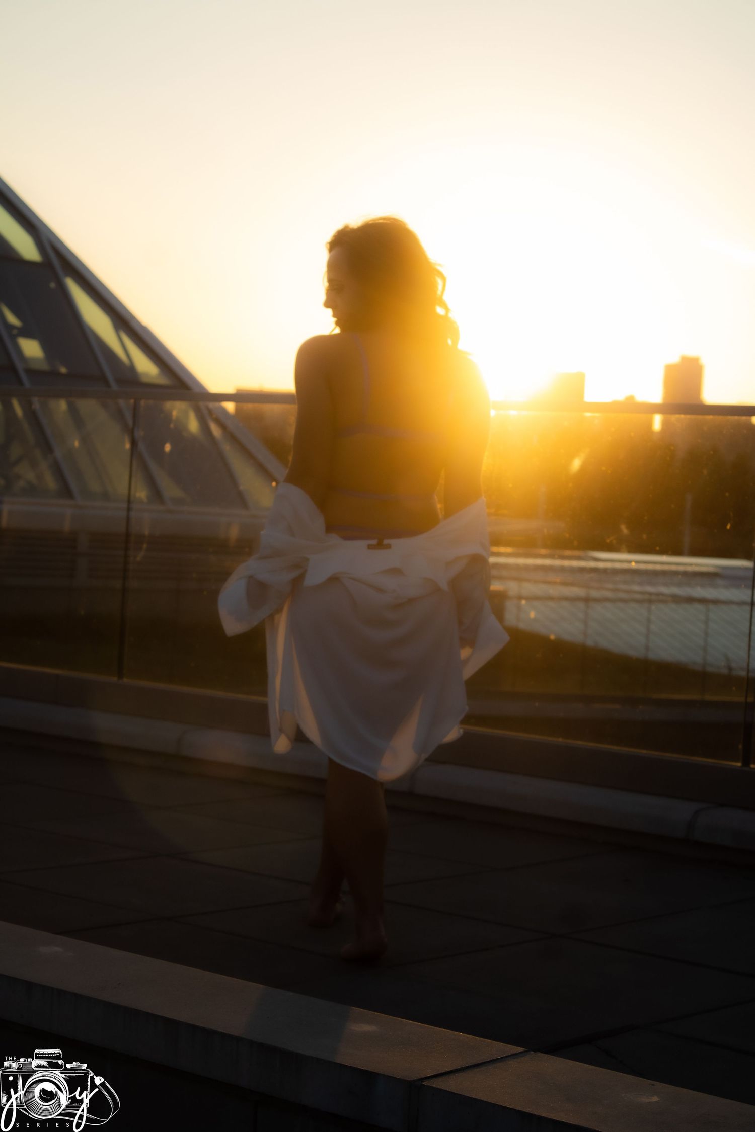 A silhouetted figure wearing a white dress stands on a rooftop against a vibrant sunset in an urban setting.