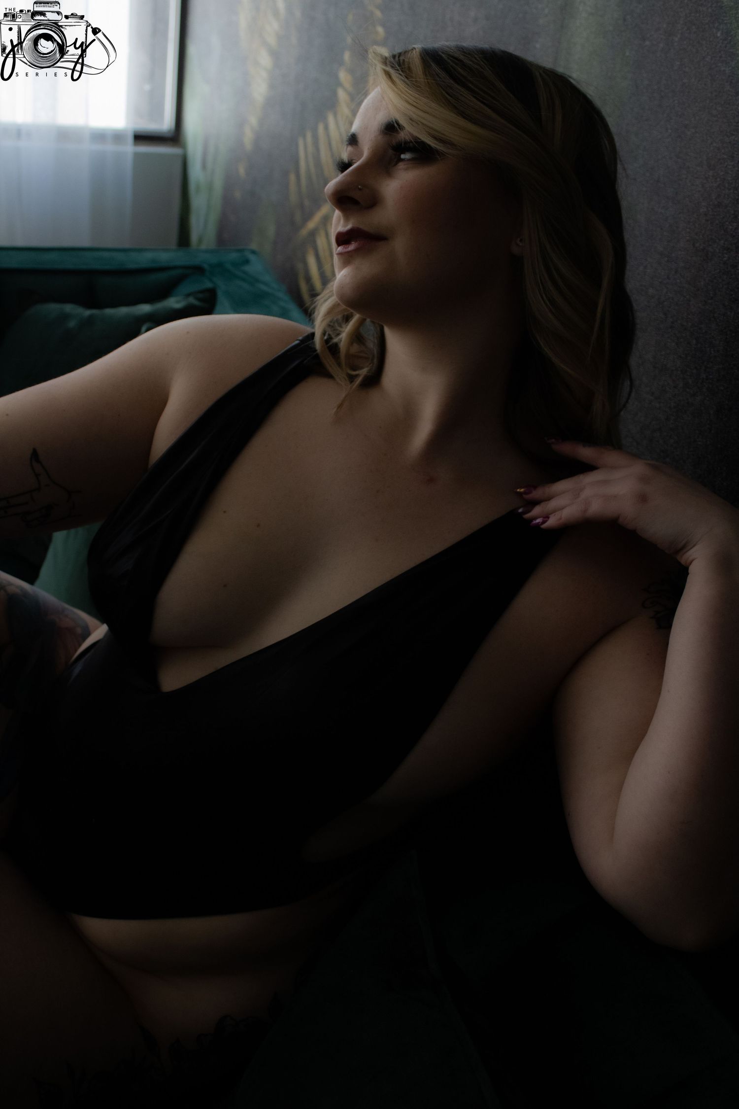 Moody boudoir portrait of someone reclining on a teal couch in dark lighting wearing black lingerie.