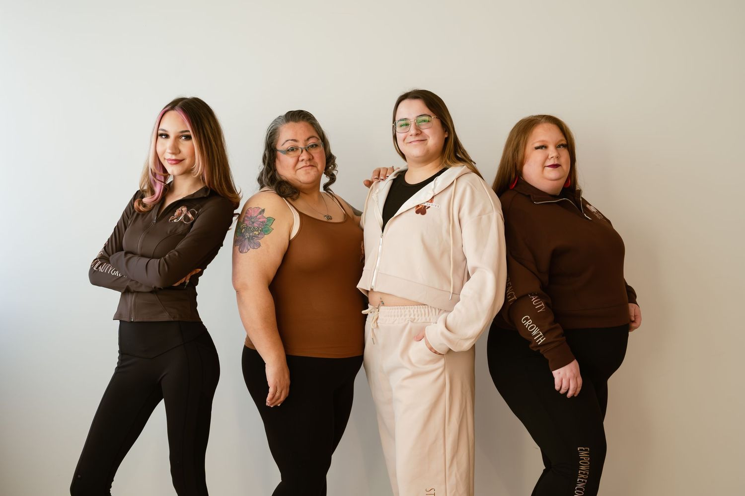 Four women posing together against a white wall, wearing casual brown and neutral outfits in a group portrait.