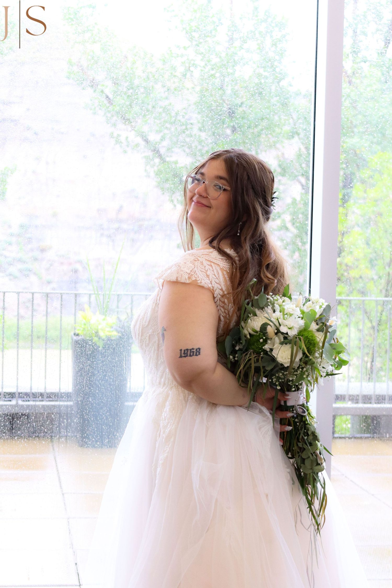 A person in a flowing white wedding dress poses gracefully near a window with a fresh floral bouquet.