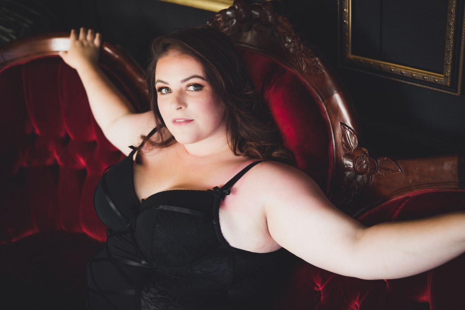 Woman in black lingerie reclining on a red velvet chair in a dramatic, moody boudoir setting.