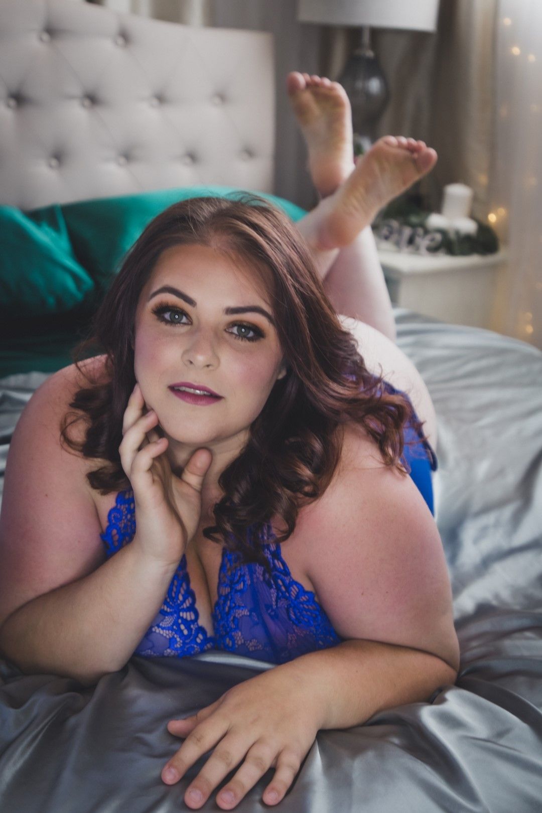 Woman in blue lace lingerie posing on a silver satin bed in a softly lit bedroom.