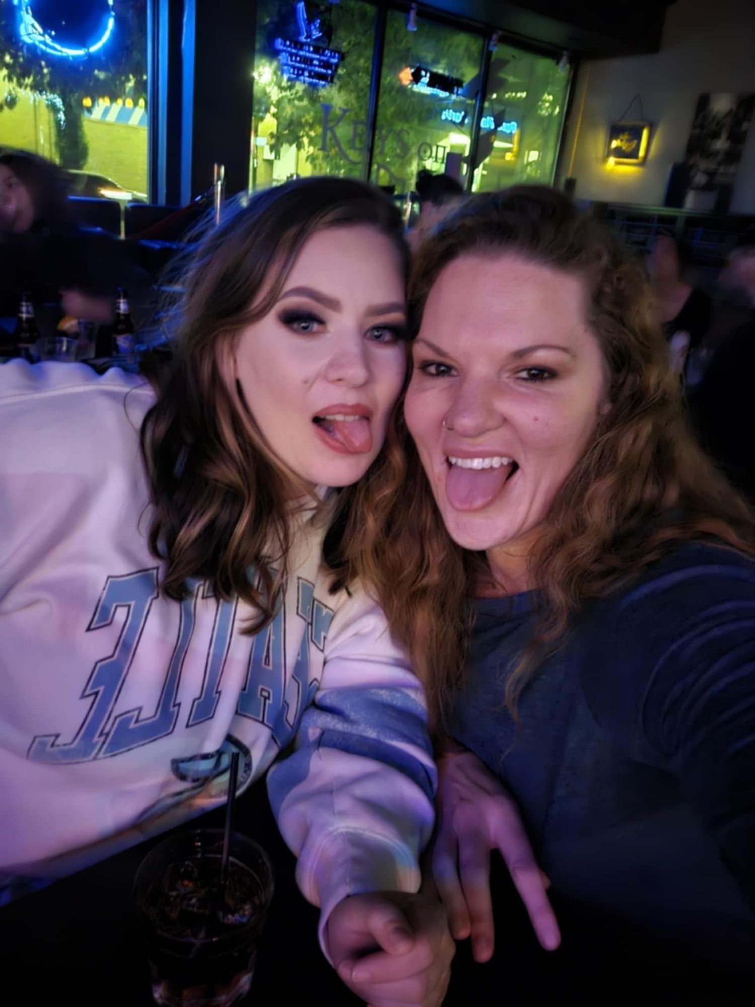 Two friends taking a fun selfie together at a bar with blue neon lights visible in the background.
