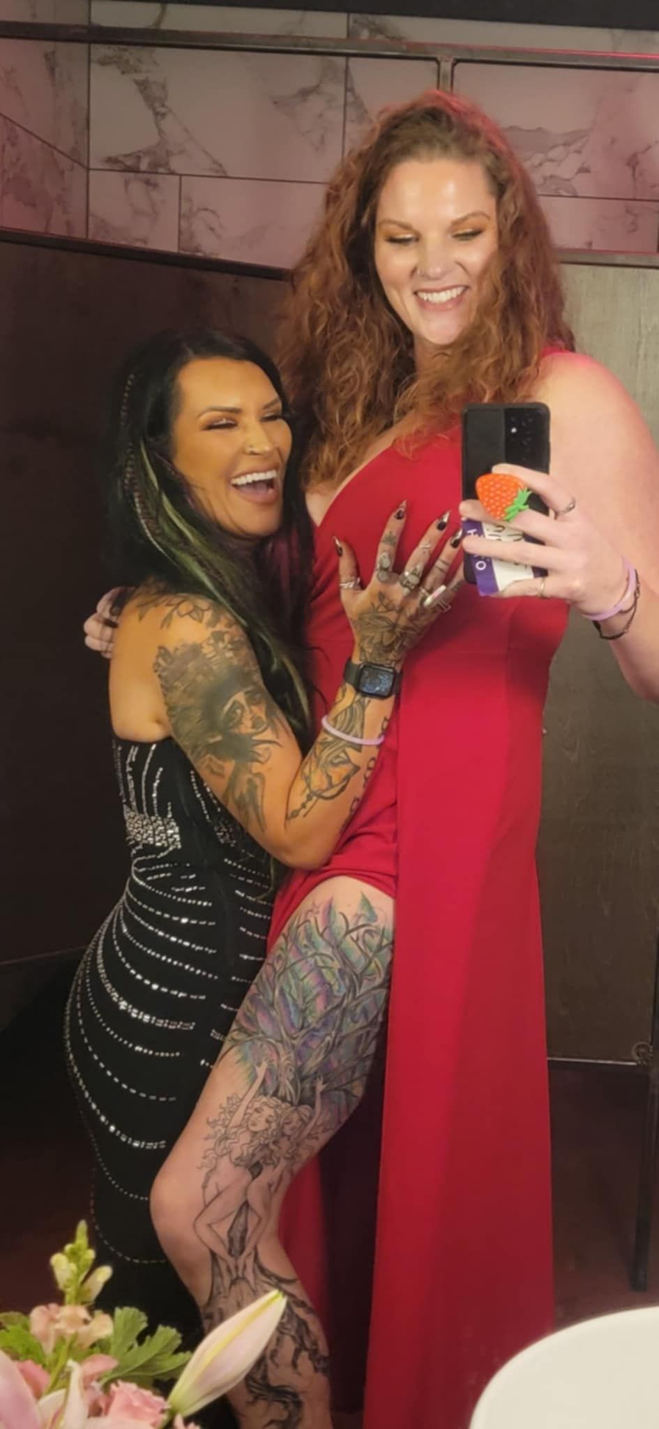 Two friends in formal dresses, one in black with tattoos and one in red, laughing while taking a selfie at an event.