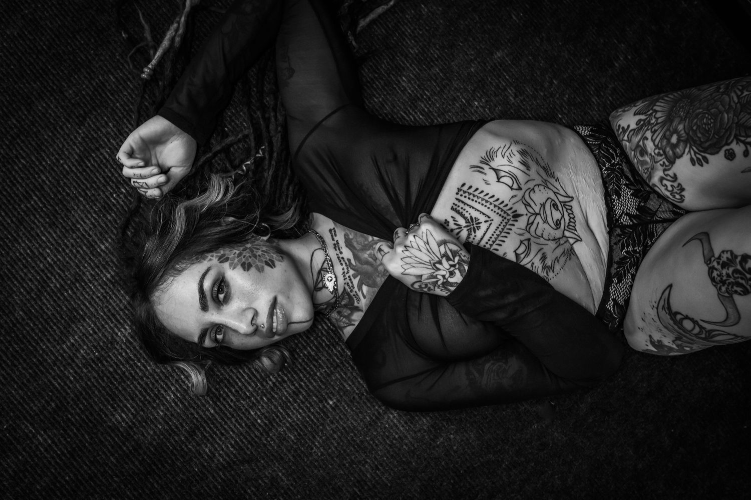 Black and white photo of a heavily tattooed woman lying on the ground wearing a dark outfit.