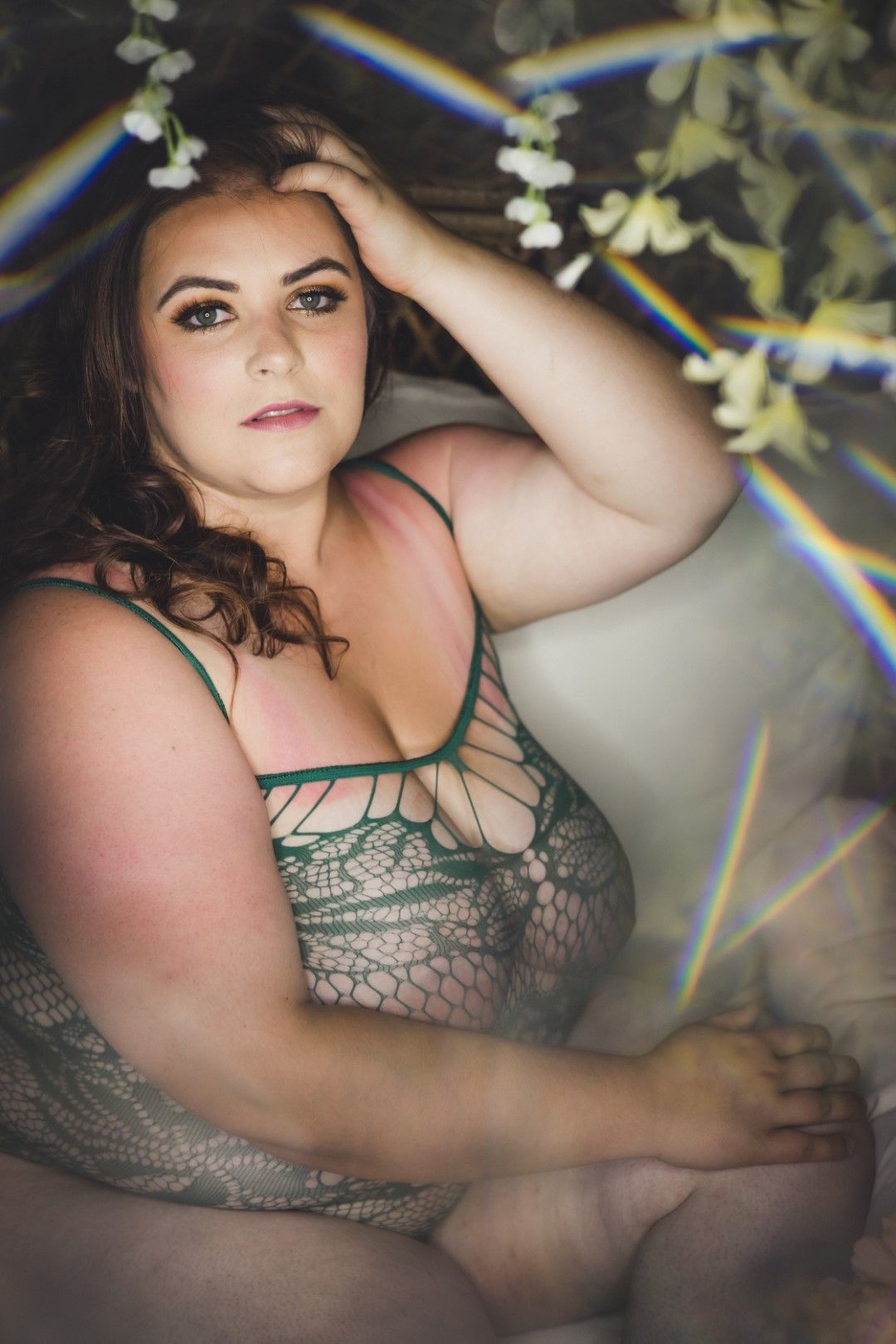 A plus-size woman in green lace lingerie poses gracefully among delicate white flowers in a boudoir setting.