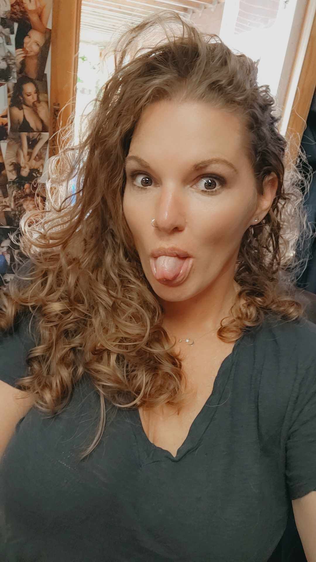 A selfie showing someone with curly brown hair wearing a black v-neck shirt making a playful expression with tongue out.