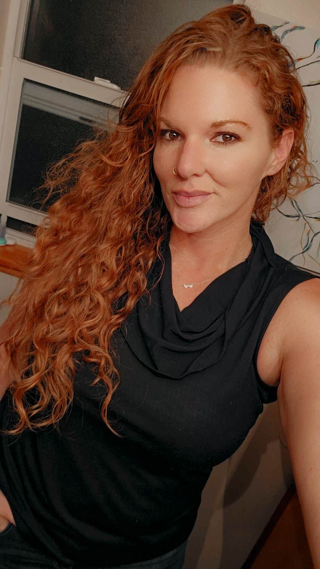 A selfie featuring long curly red hair, a black sleeveless top with cowl neck, and delicate necklace.