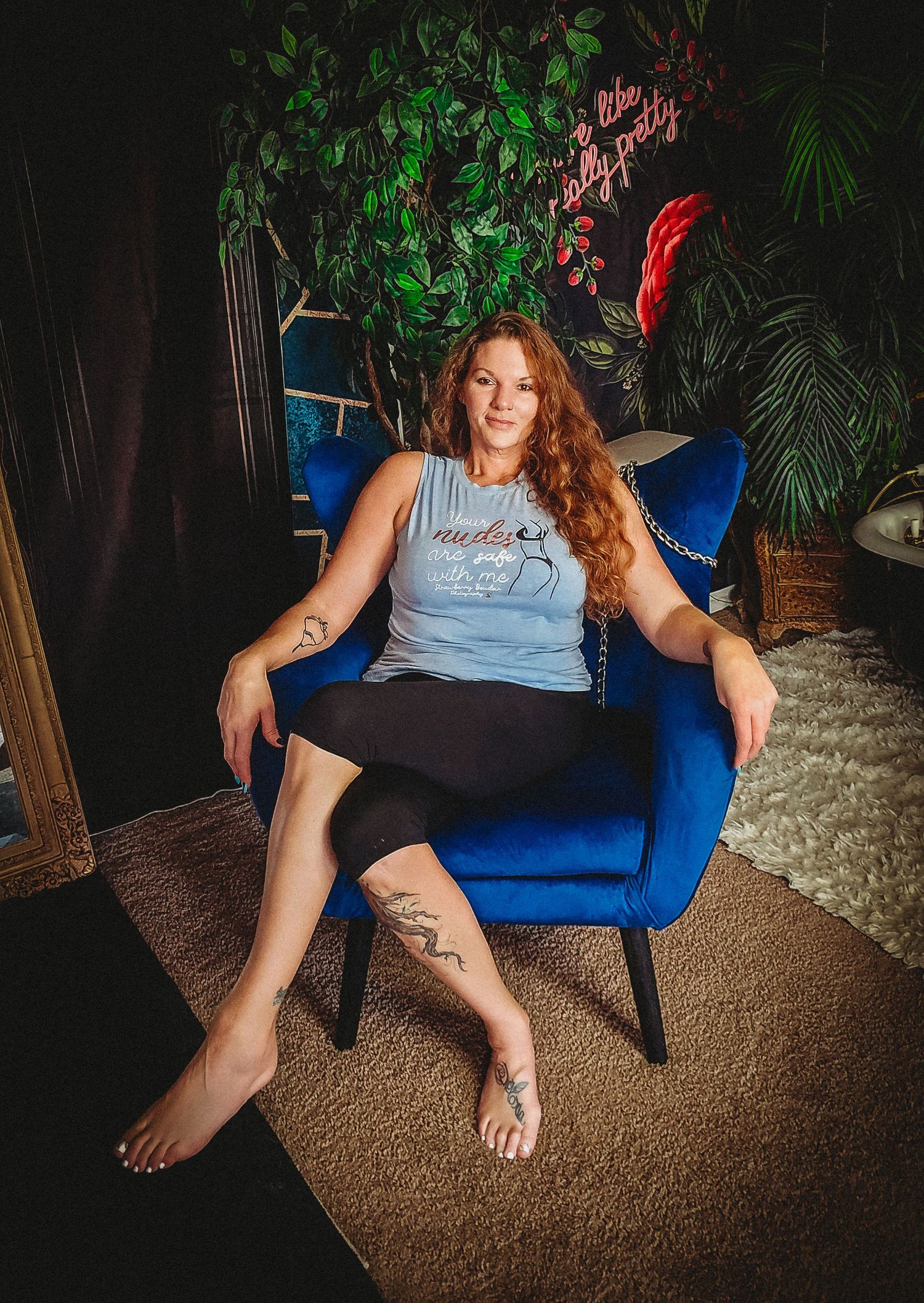 Woman with red hair and tattoos sitting in a blue velvet chair surrounded by lush green plants.
